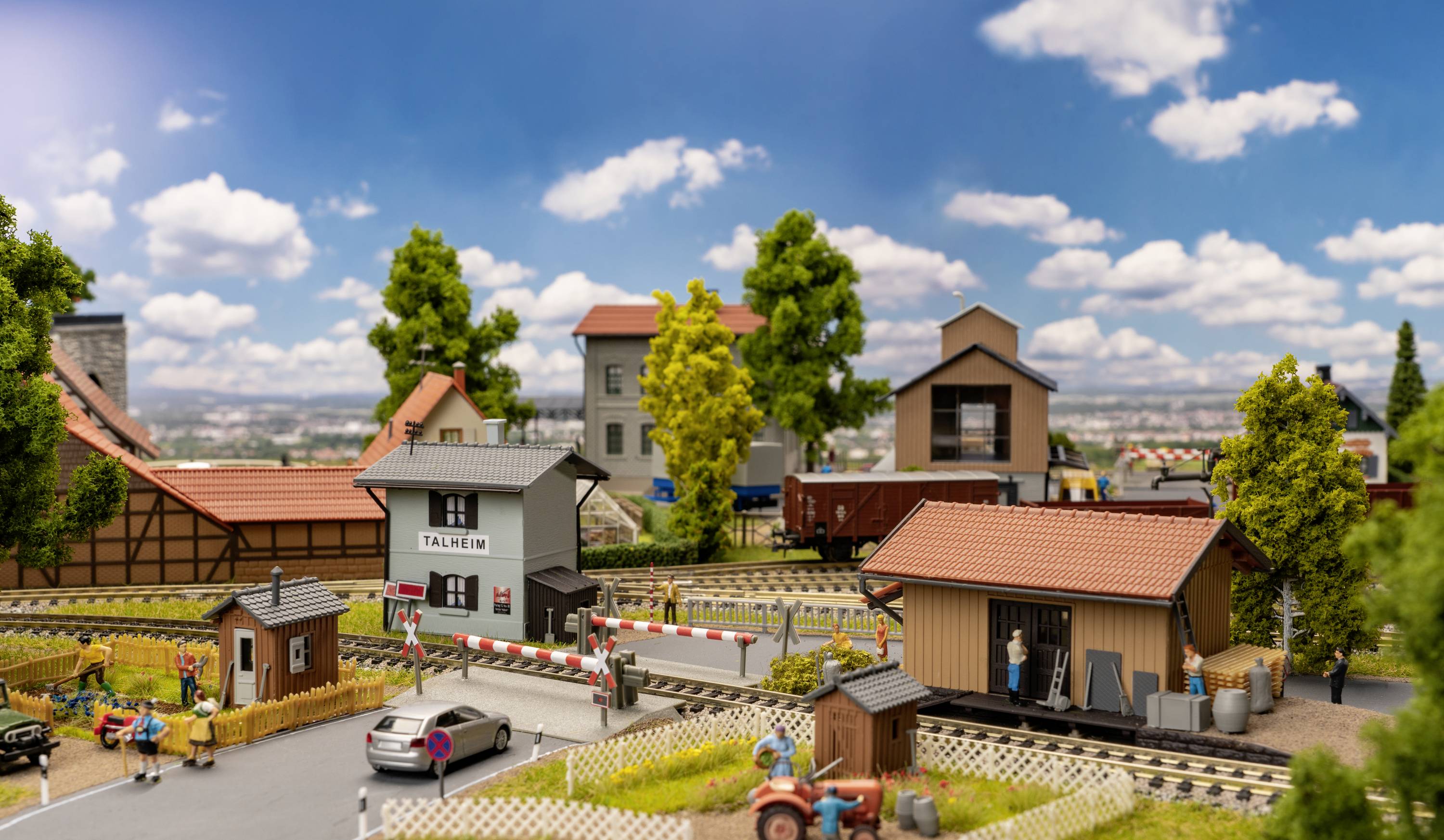 Modellbahnlandschaft mit kleinen Gebäuden, Gleisen, Autos und Bäumen unter blauem Himmel mit Wolken. Menschenfiguren beleben die Szene.
