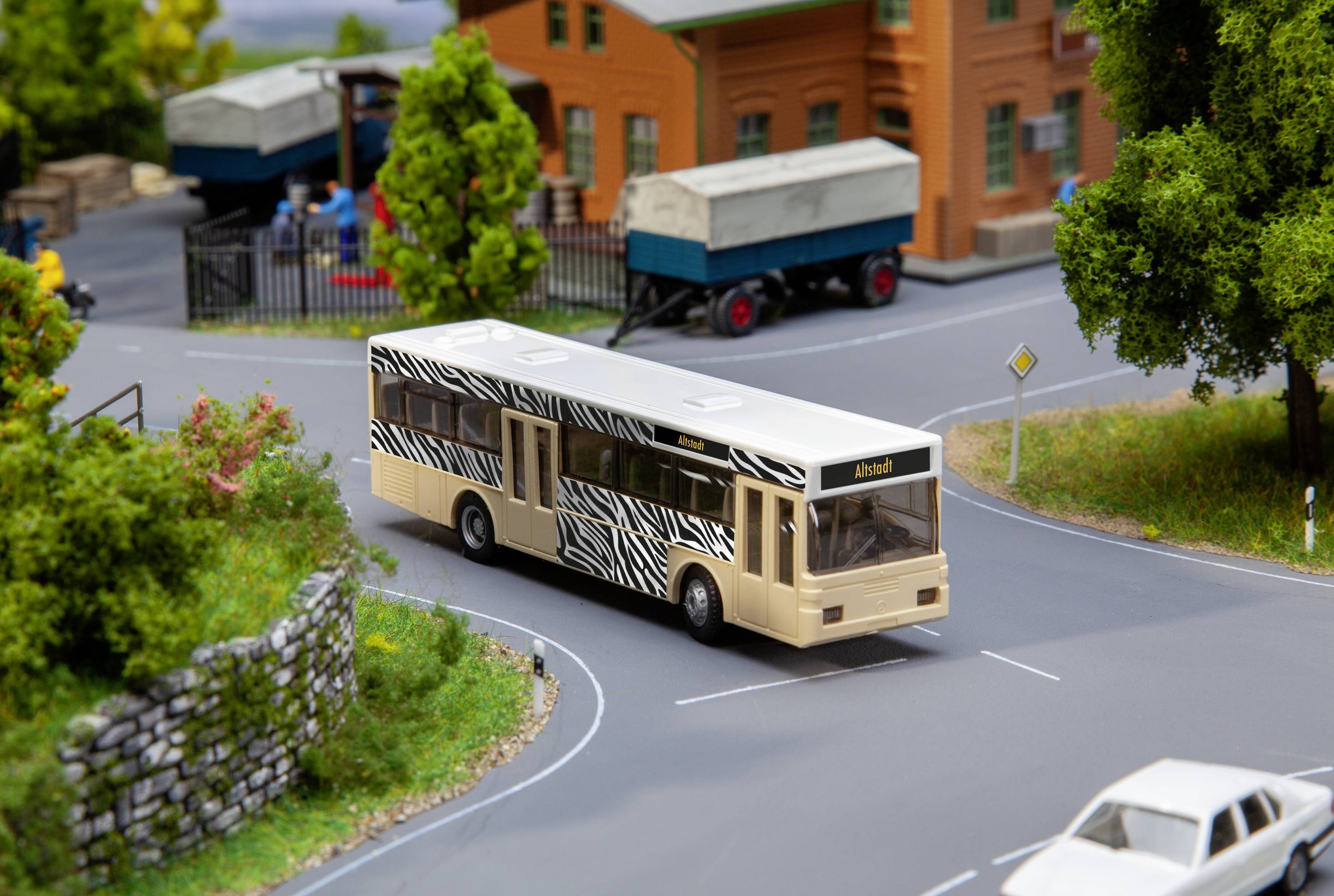 Ein Modellbus mit Zebramuster fährt auf einer Miniaturstraße. Im Hintergrund sind Bäume, ein Zaun und ein Lastwagen sichtbar.