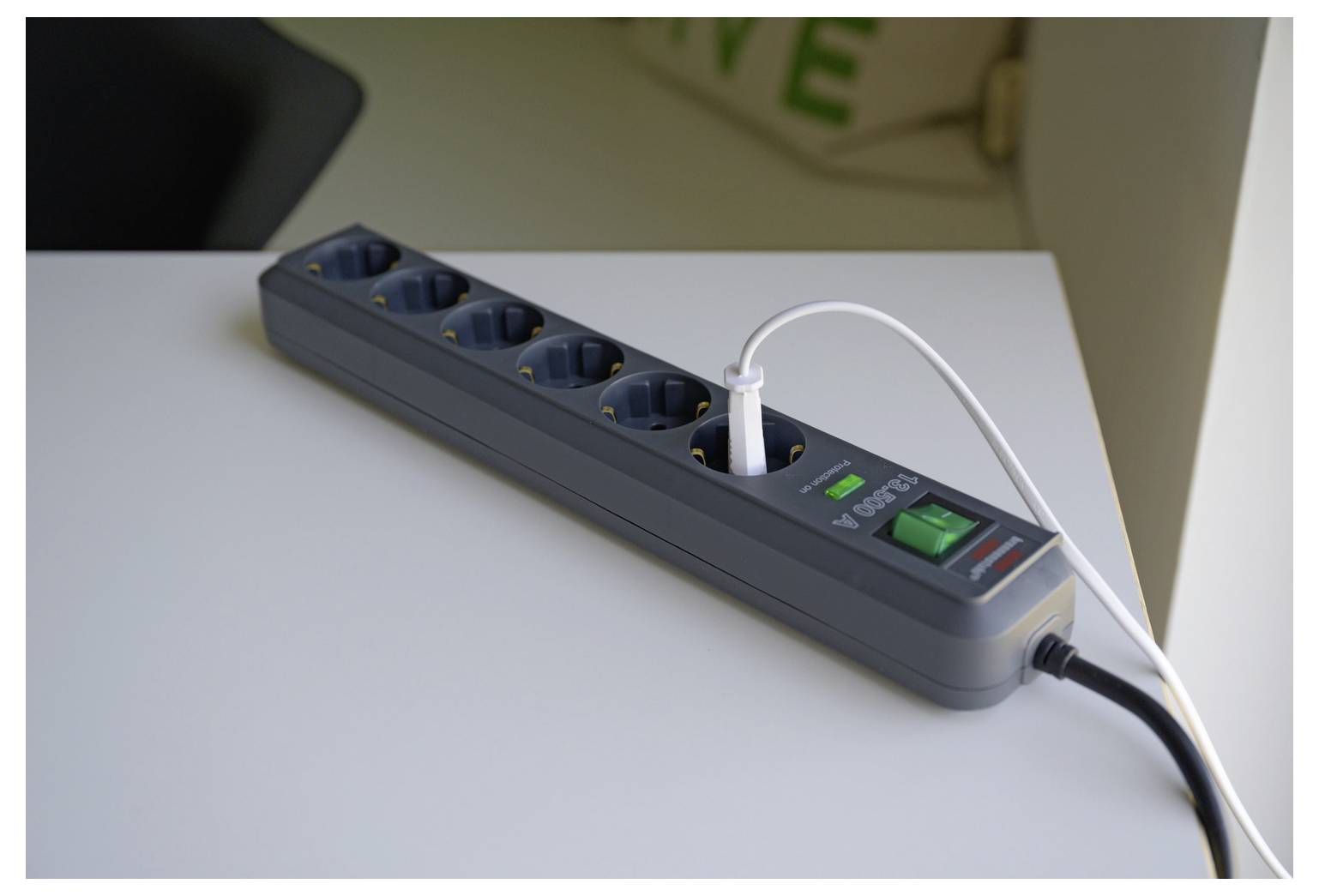 Ein Steckerleisten mit mehreren Steckdosen steht auf einem Schreibtisch, wobei ein weißes Kabel eingesteckt ist und ein grüner Ein-/Ausschalter sichtbar ist.