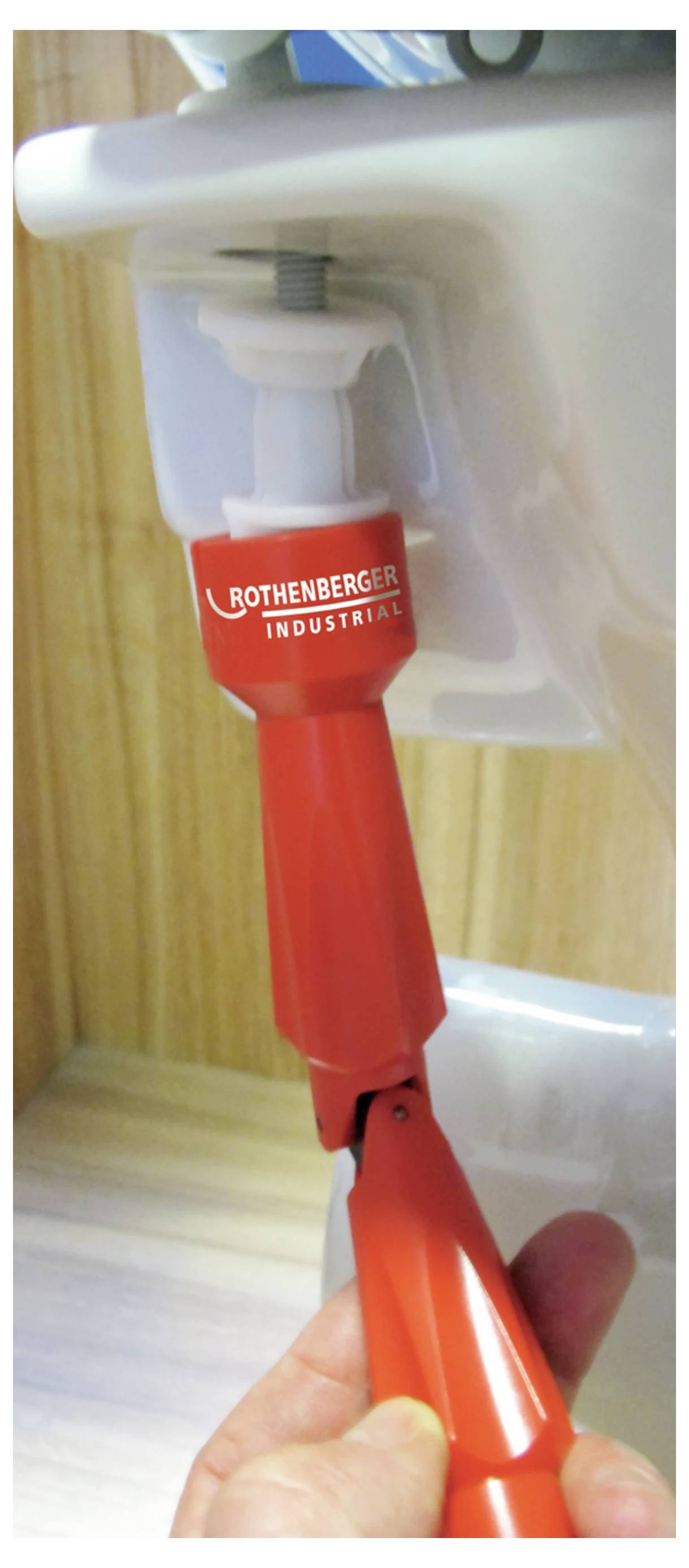 Ein rotes Klempnerwerkzeug wird verwendet, um eine Schraube unter einem Toilettentank festzuziehen. Das Werkzeug ist mit der Marke 'Rothenberger Industrial' gekennzeichnet.