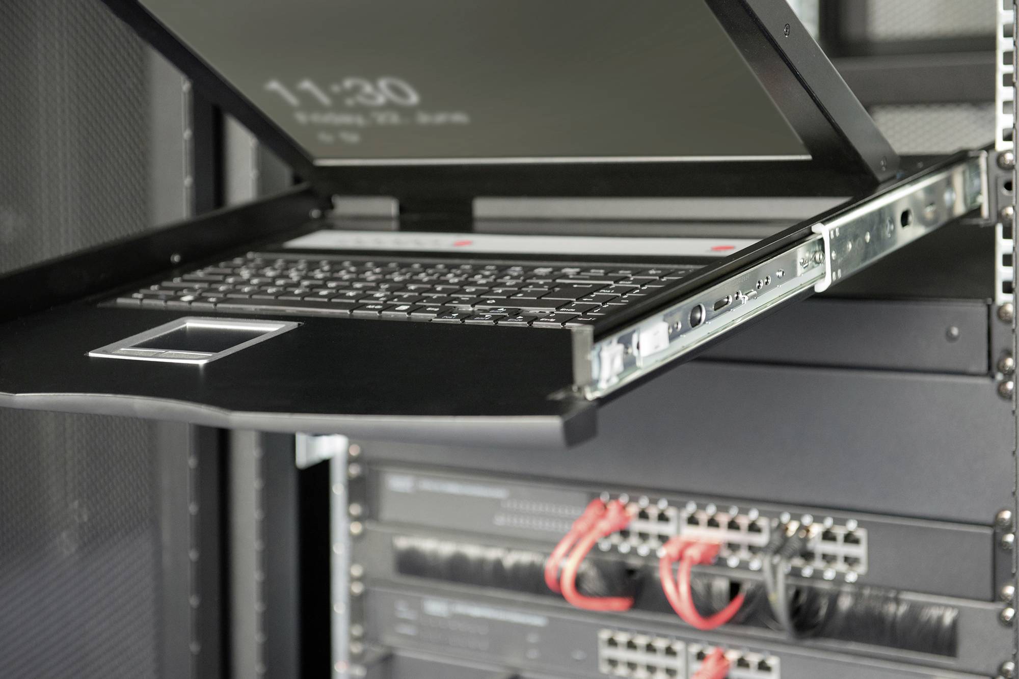 Ein geöffneter Laptop in einem Server-Rack zeigt eine Uhrzeit auf dem Bildschirm. Darunter sind Netzwerk-Switches und Kabel zu sehen.