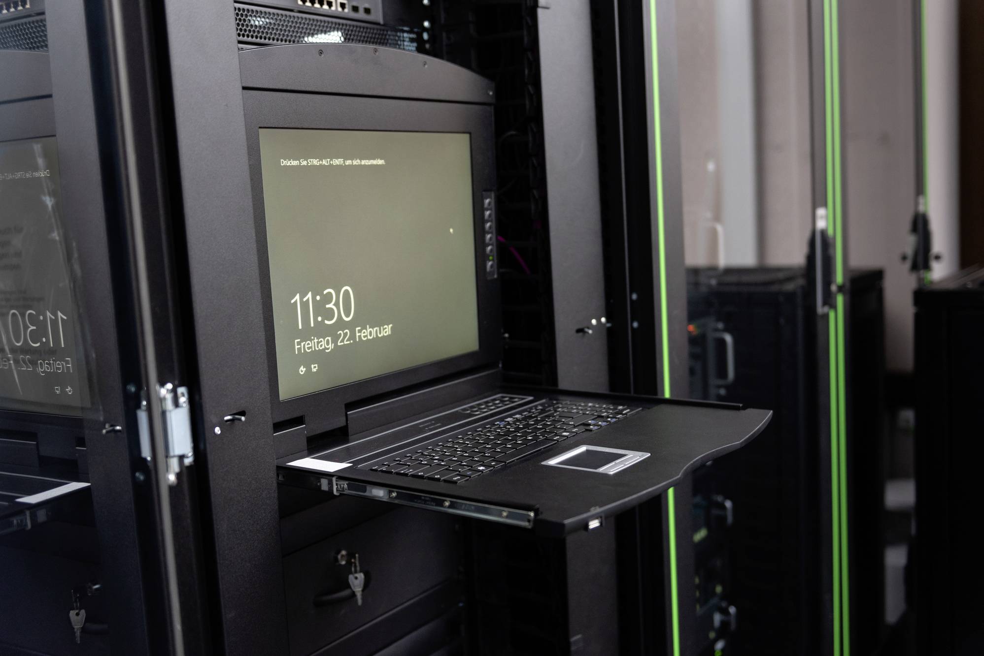 Ein Serverraum mit einem aufgeklappten Laptop in einem Serverschrank. Der Bildschirm zeigt das Datum Freitag, 22. Februar und die Uhrzeit 11:30.