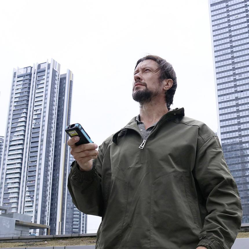 Ein Mann im Freien, der ein Smartphone hält, mit Wolkenkratzern im Hintergrund. Er trägt eine khakifarbene Jacke.