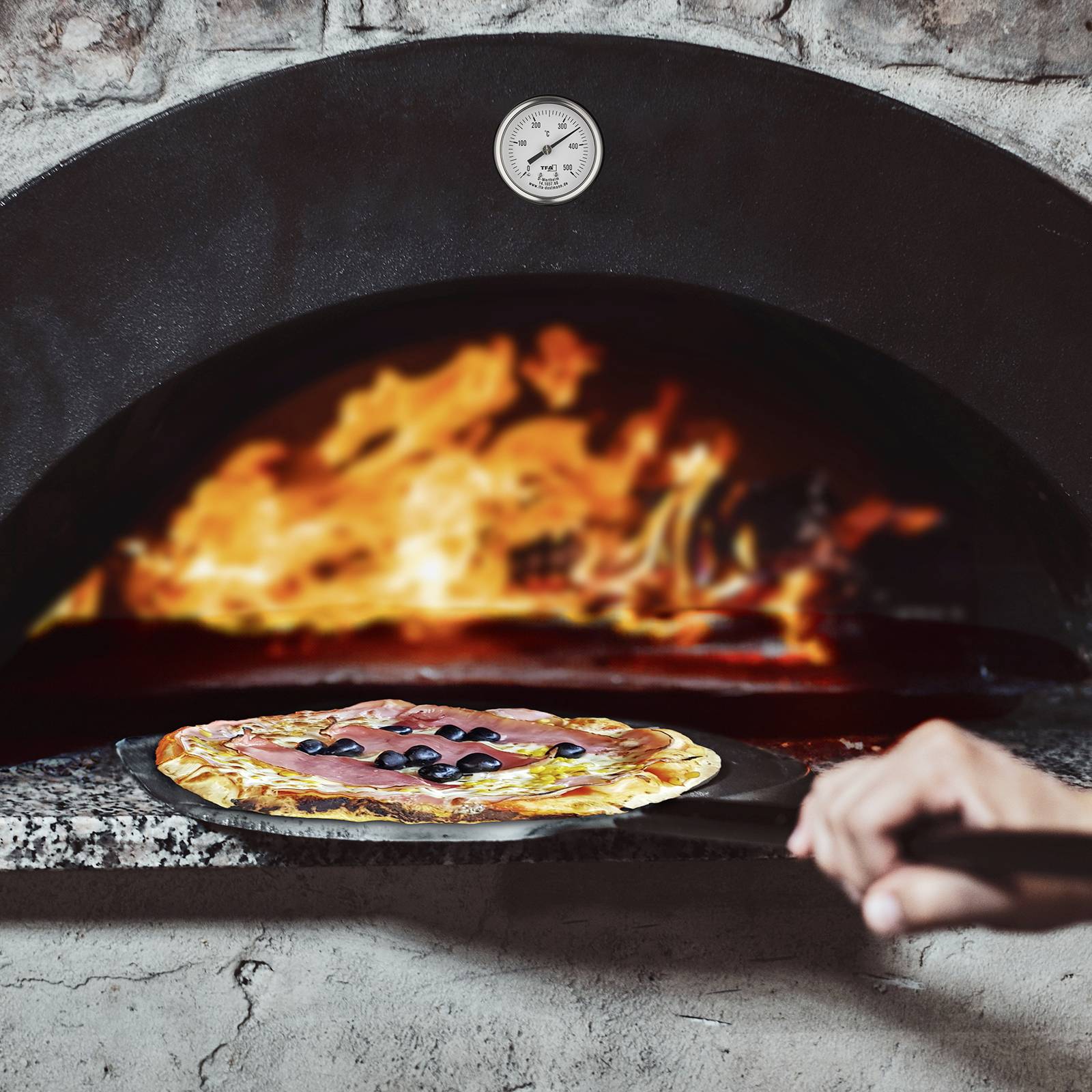 Eine Hand hält einen Pizzaheber mit einer Pizza vor einem traditionellen steinernen Holzofen, in dem ein Feuer brennt.