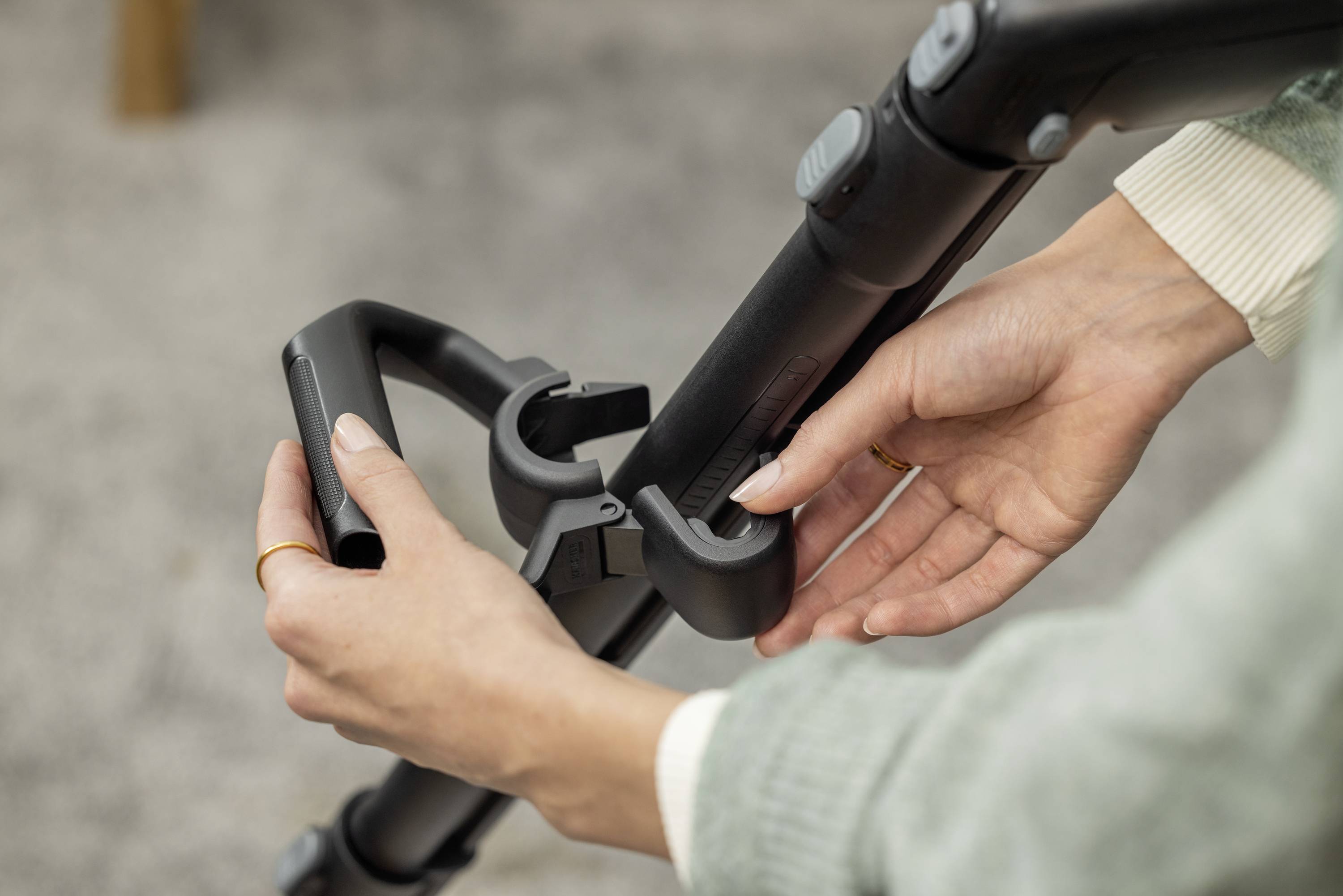 Eine Person befestigt ein schwarzes Plastikzubehör an einem Stangengriff eines Kinderwagens. Hände justieren die Verbindung.