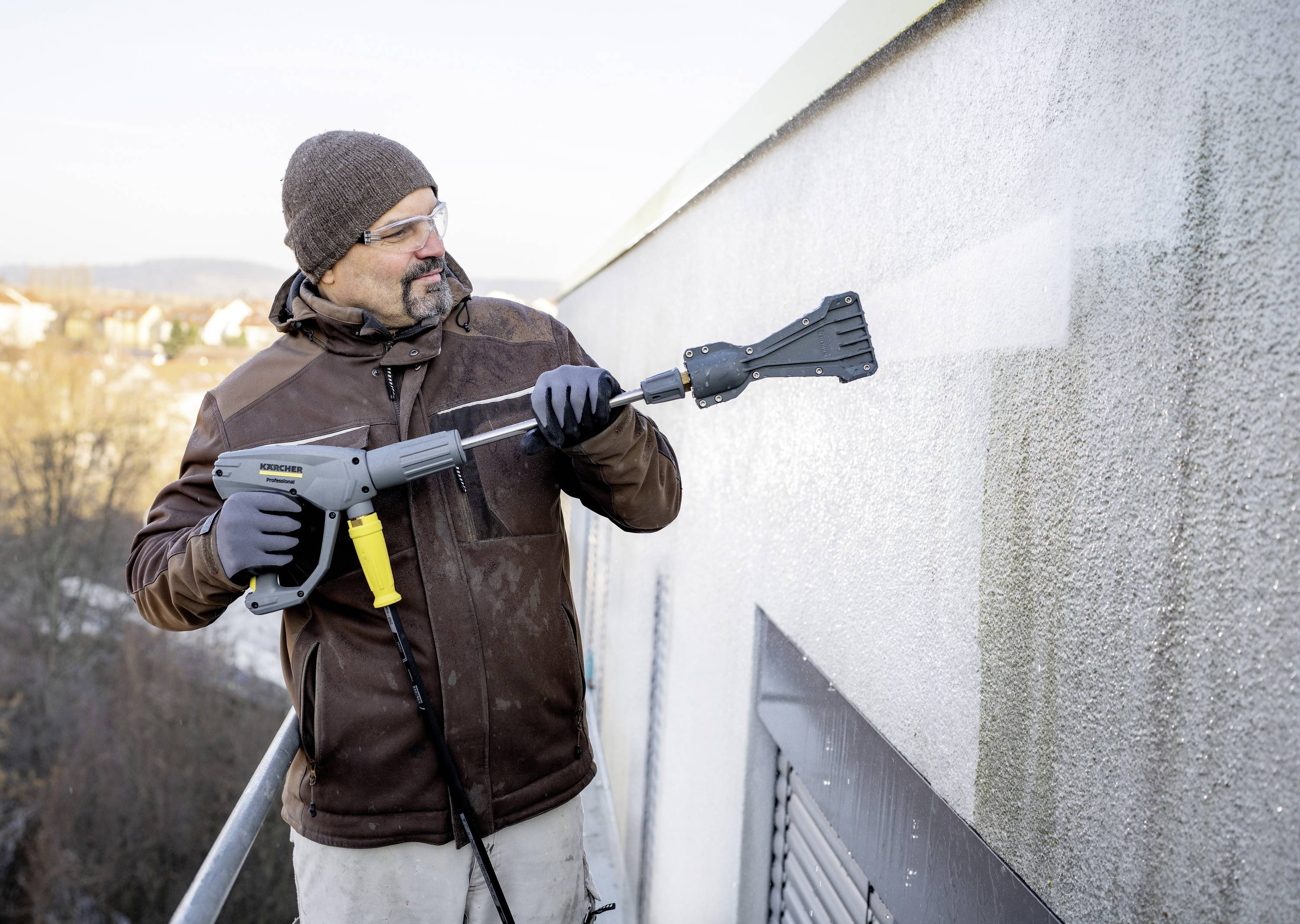 Mann reinigt Hausfassade mit Hochdruckreiniger. Trägt Mütze und Schutzbrille. Gebäude im Hintergrund bei klarer Witterung.