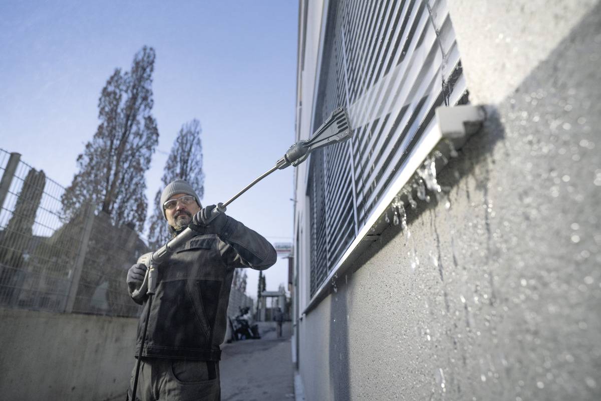 Eine Person in warmer Kleidung reinigt die Außenfensterjalousien eines Gebäudes mit einer langen Bürste, wobei Wasser spritzt, an einem sonnigen Tag.