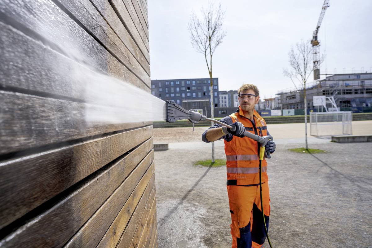 Eine Person in orangefarbener Sicherheitskleidung reinigt eine Holzwand mit einem Hochdruckreiniger auf einer Baustelle, wobei Gebäude und ein Kran im Hintergrund zu sehen sind.