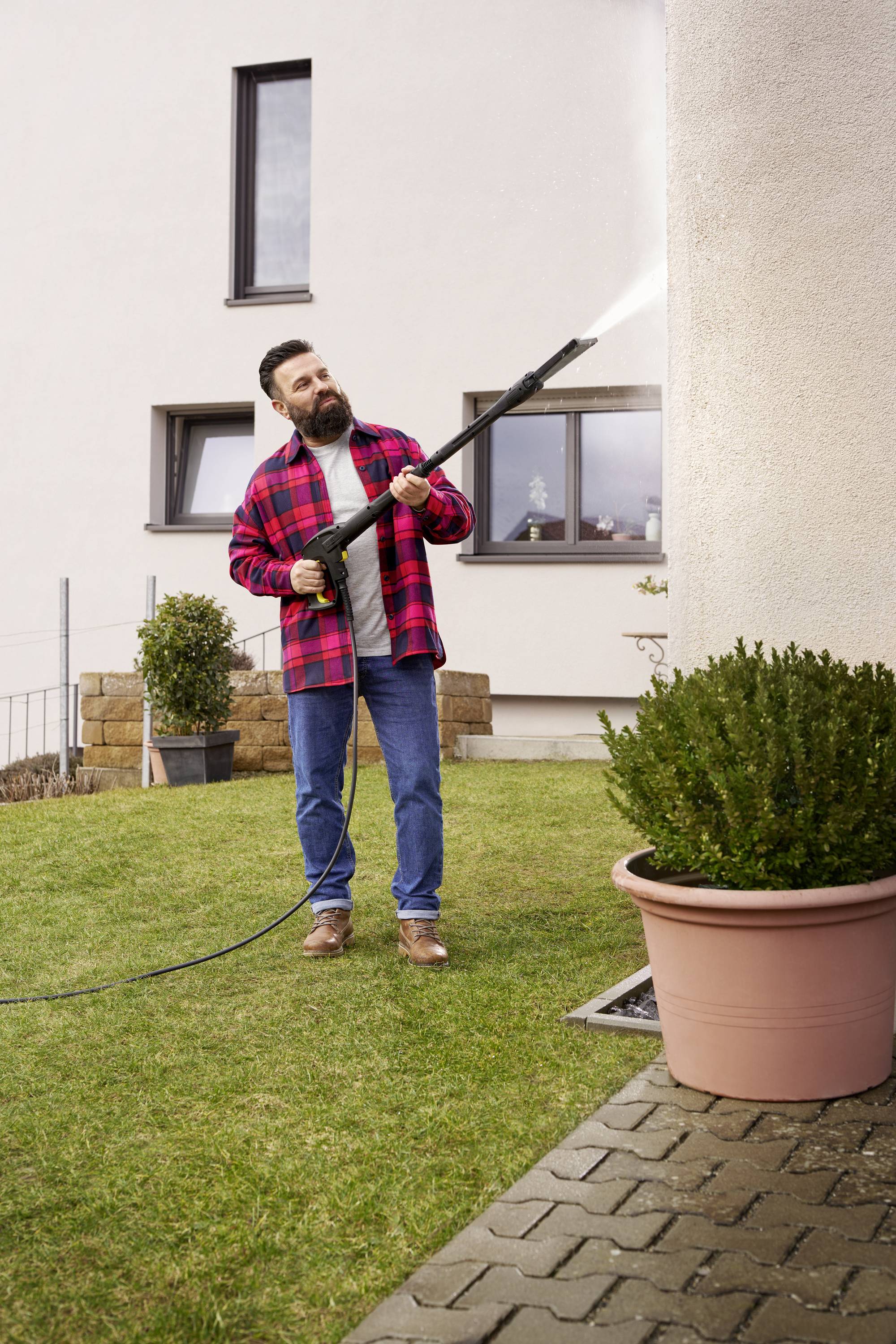 Ein Mann reinigt mit einem Hochdruckreiniger die Fassade eines Hauses, umgeben von einem Garten mit Pflanzkübeln und Rasen.