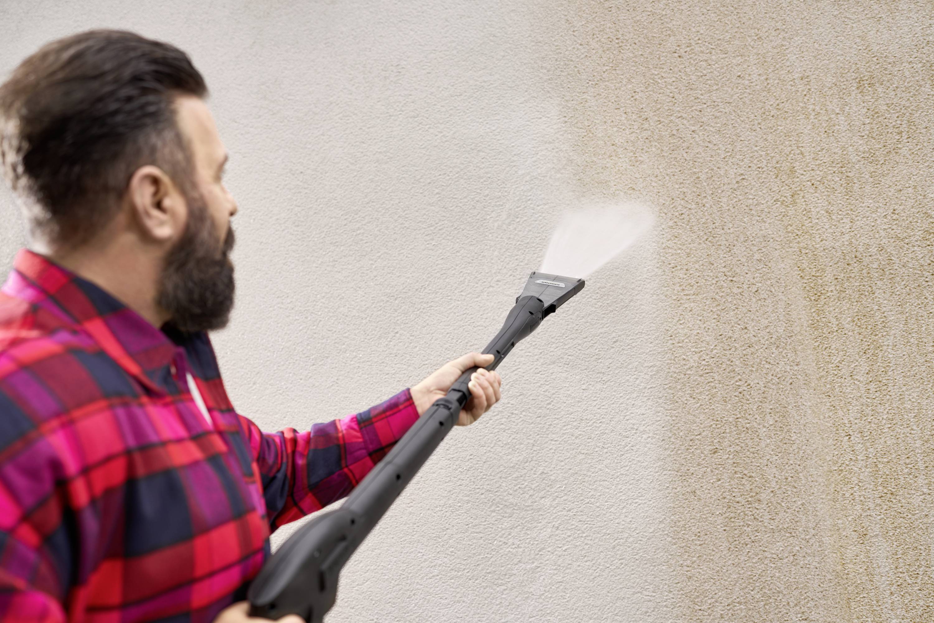 Eine Person in einem karierten Hemd reinigt eine verschmutzte Wand mit einem Hochdruckreiniger. Die Wand wird sichtbar sauberer.