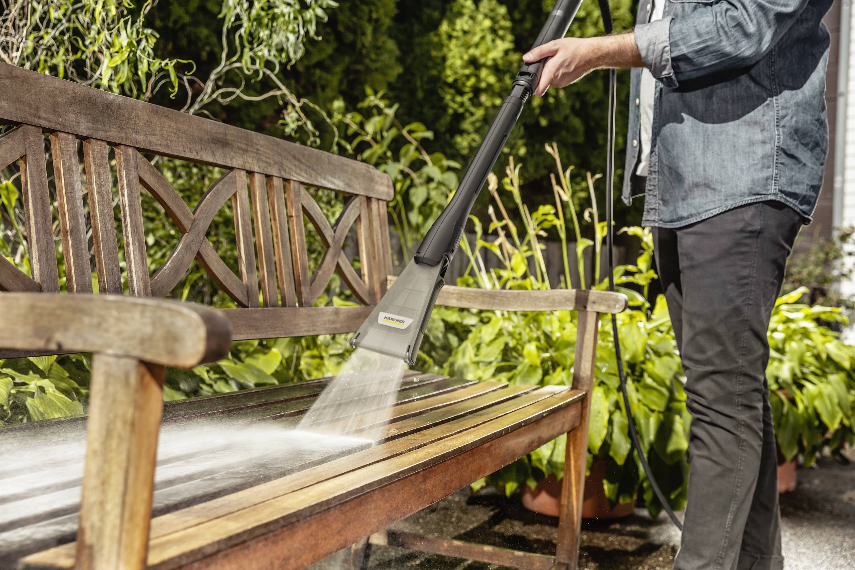 Eine Person reinigt eine Holzbank im Garten mit einem Hochdruckreiniger. Pflanzen und Bäume sind im Hintergrund sichtbar.