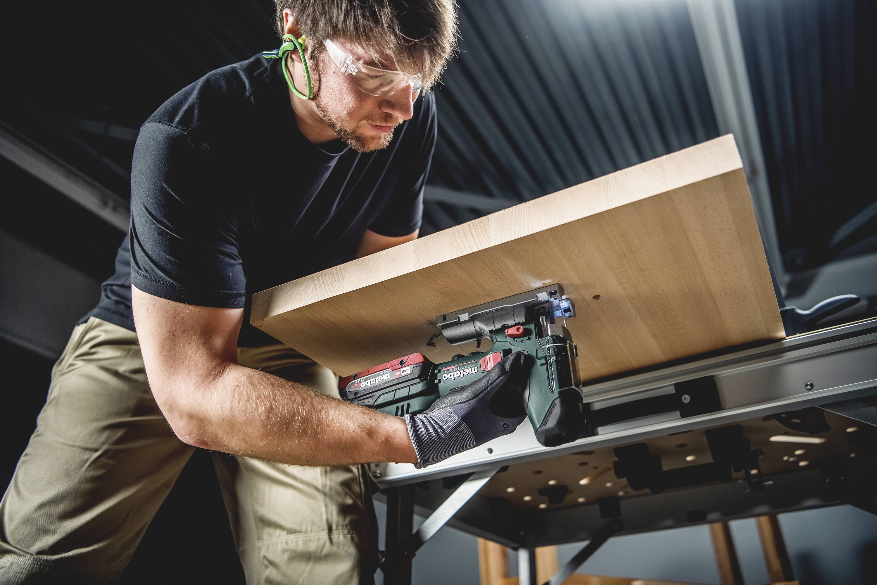 Ein Mann befestigt eine Holzplatte mit einem Elektrowerkzeug auf einem Arbeitstisch. Er trägt Sicherheitsbrille und Handschuhe.