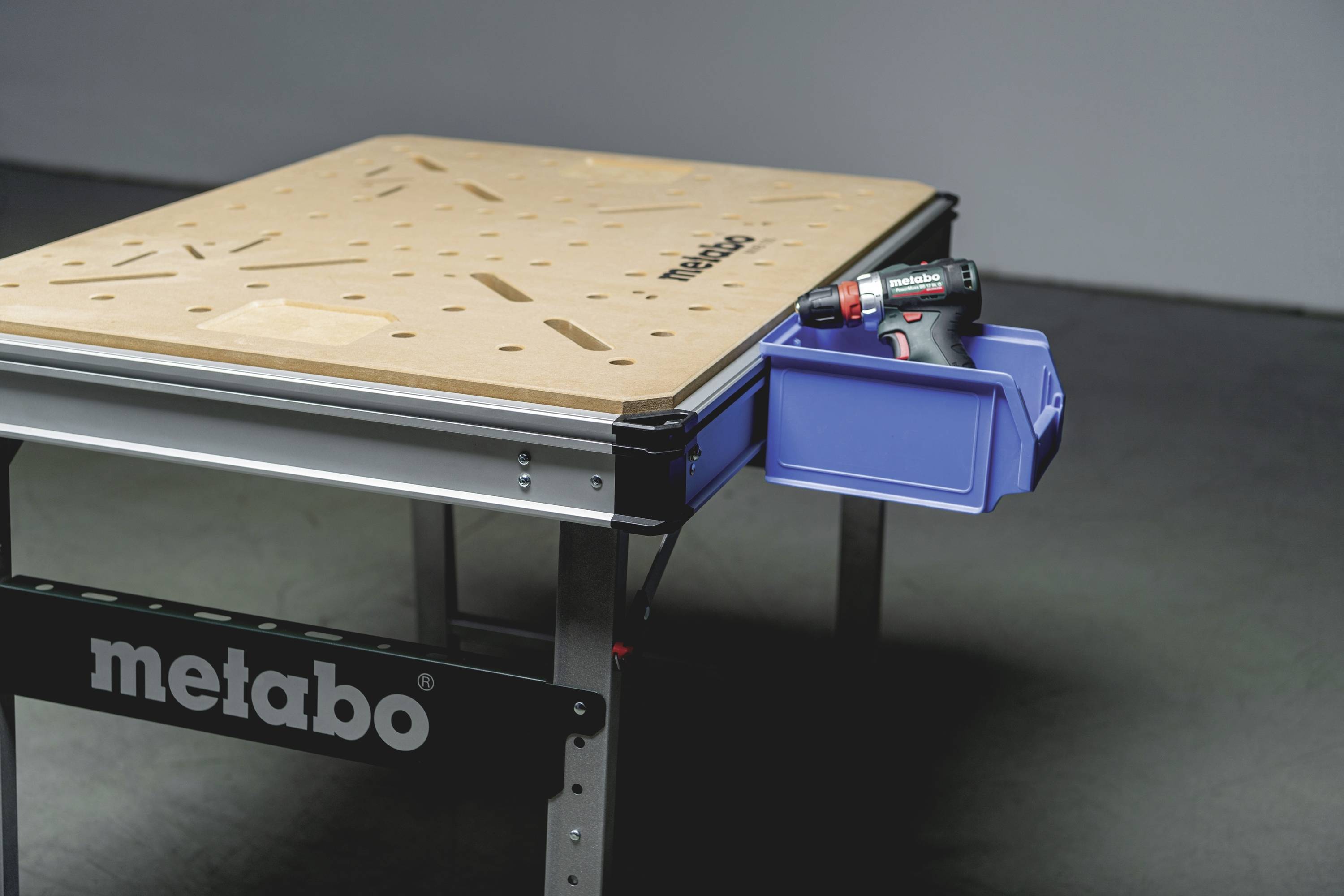 Ein Werkstatttisch mit einem perforierten Holzbrett, das von 'metabo' gekennzeichnet ist. Rechts hängt eine blaue Aufbewahrungsbox mit einer Bohrmaschine darin.