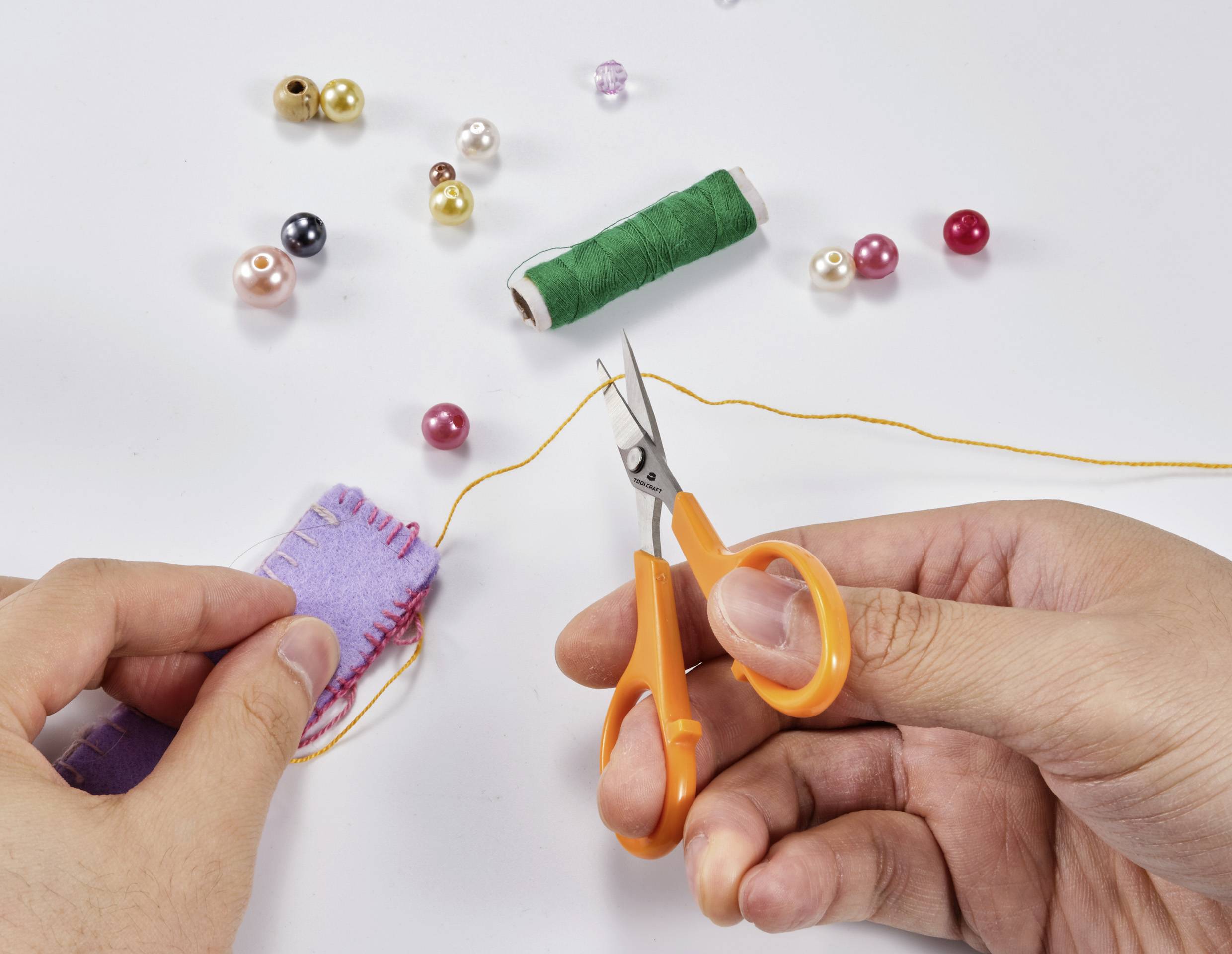 Eine Person näht mit einem lilafarbenen Filzstück, orangefarbener Schere, Nähgarn und Perlen auf hellem Hintergrund.