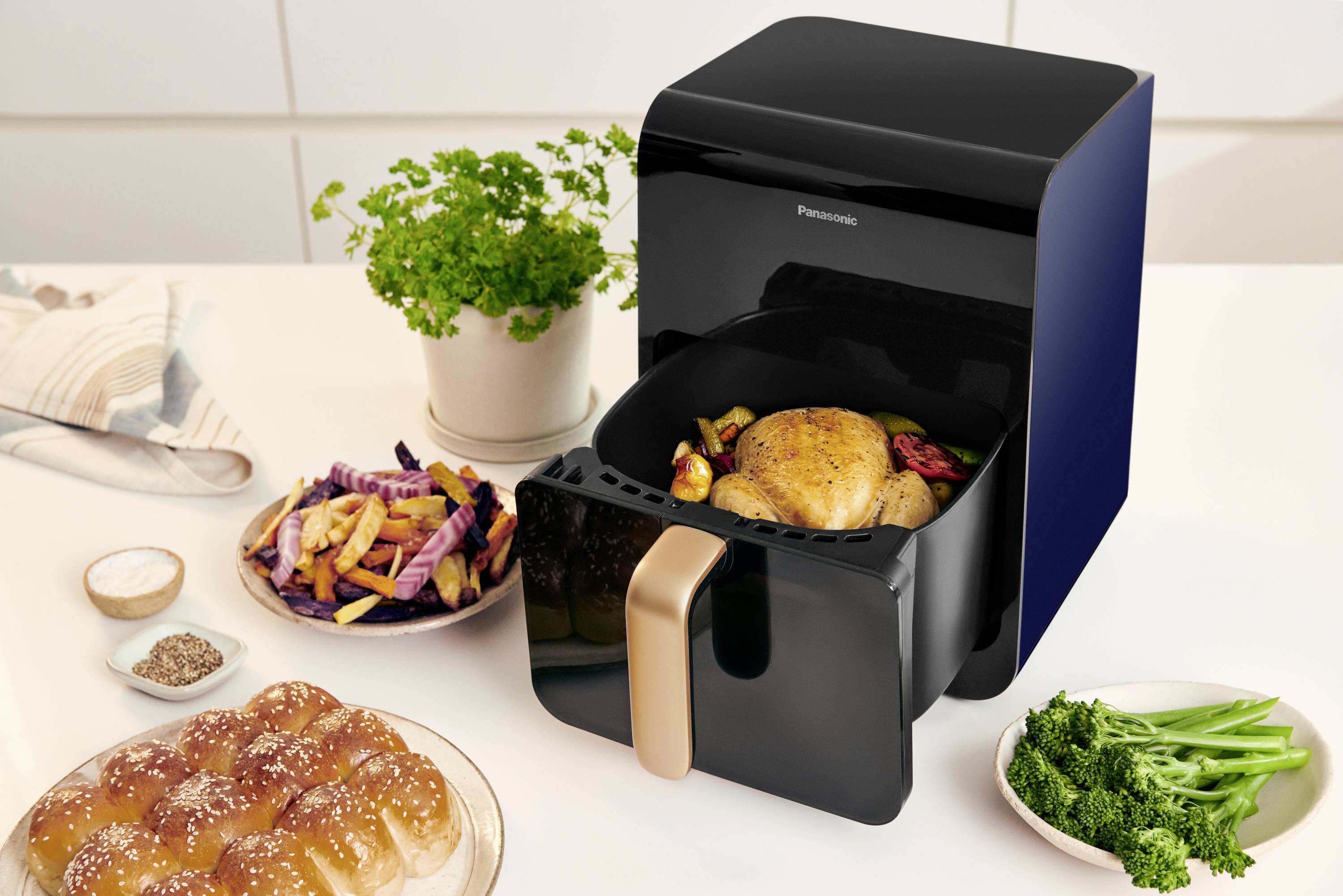 Ein schwarzer Airfryer mit goldener Griff steht auf einer Küchenarbeitsplatte. Darin ein gebratenes Hähnchen mit Gemüse. Daneben Brot, Pommes und Kräuter.