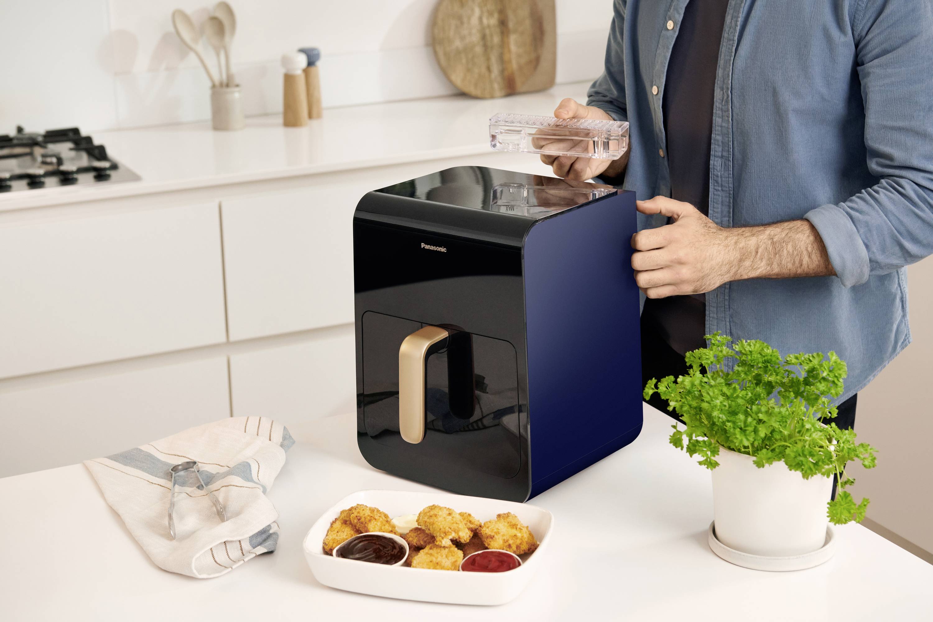 Person nutzt modernen Wasserfilter in heller Küche. Tisch zeigt Servietten, Snacks und Topf mit grüner Pflanze.