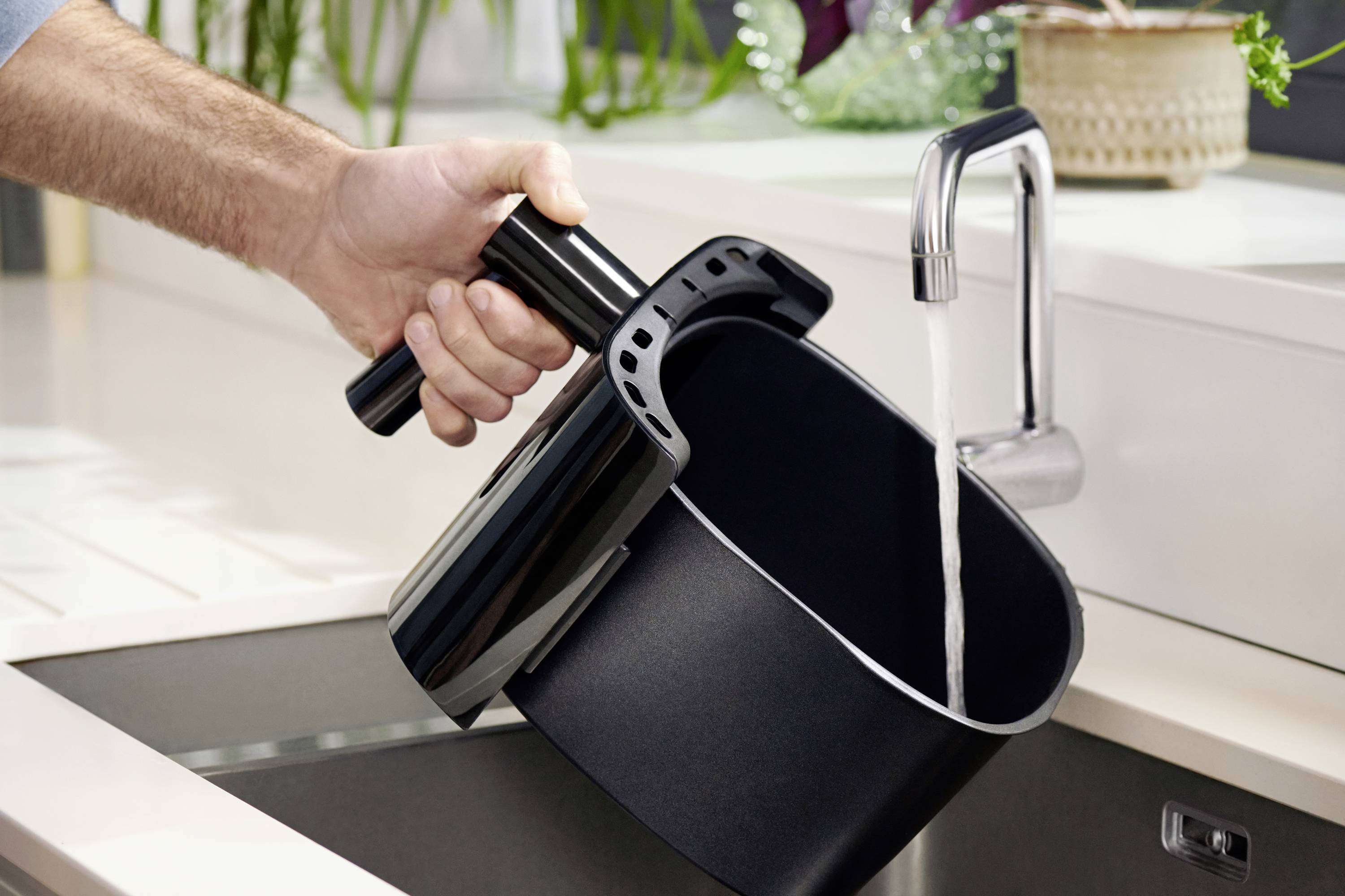 Eine Hand hält einen Airfryer-Korb, der unter einem Wasserhahn ausgespült wird.