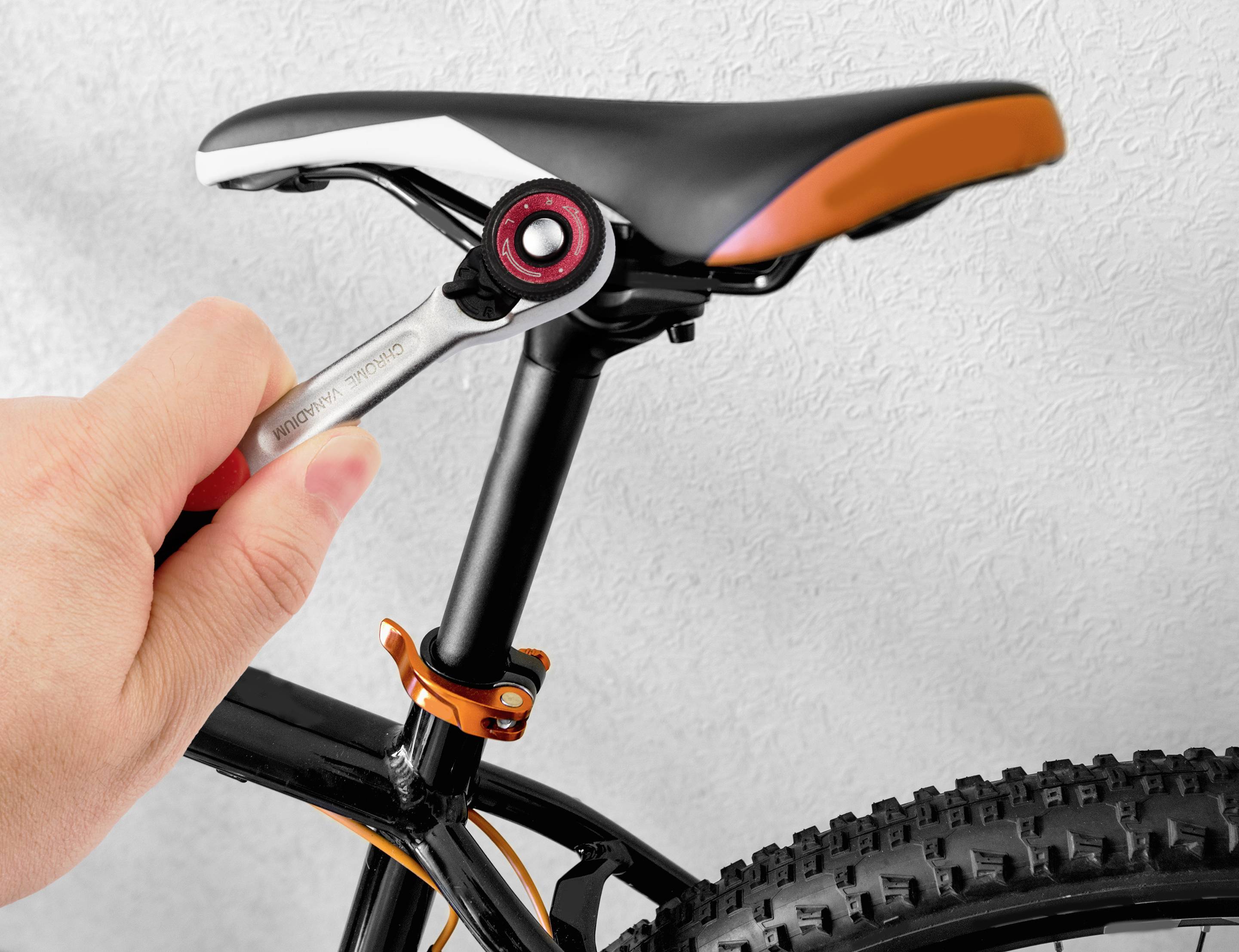 Eine Hand zieht mit einem Schraubenschlüssel die Sattelklemme eines Fahrrads fest. Das Fahrrad hat grobstollige Reifen und einen schwarzen Rahmen.