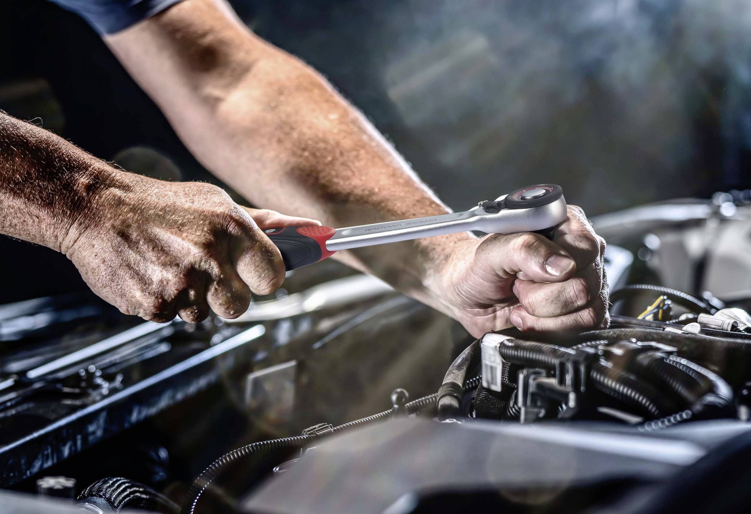 Zwei Hände reparieren einen Motorteil mit einem Schraubenschlüssel. Die Umgebung ist eine Werkstatt mit Fokus auf die Reparaturarbeit.