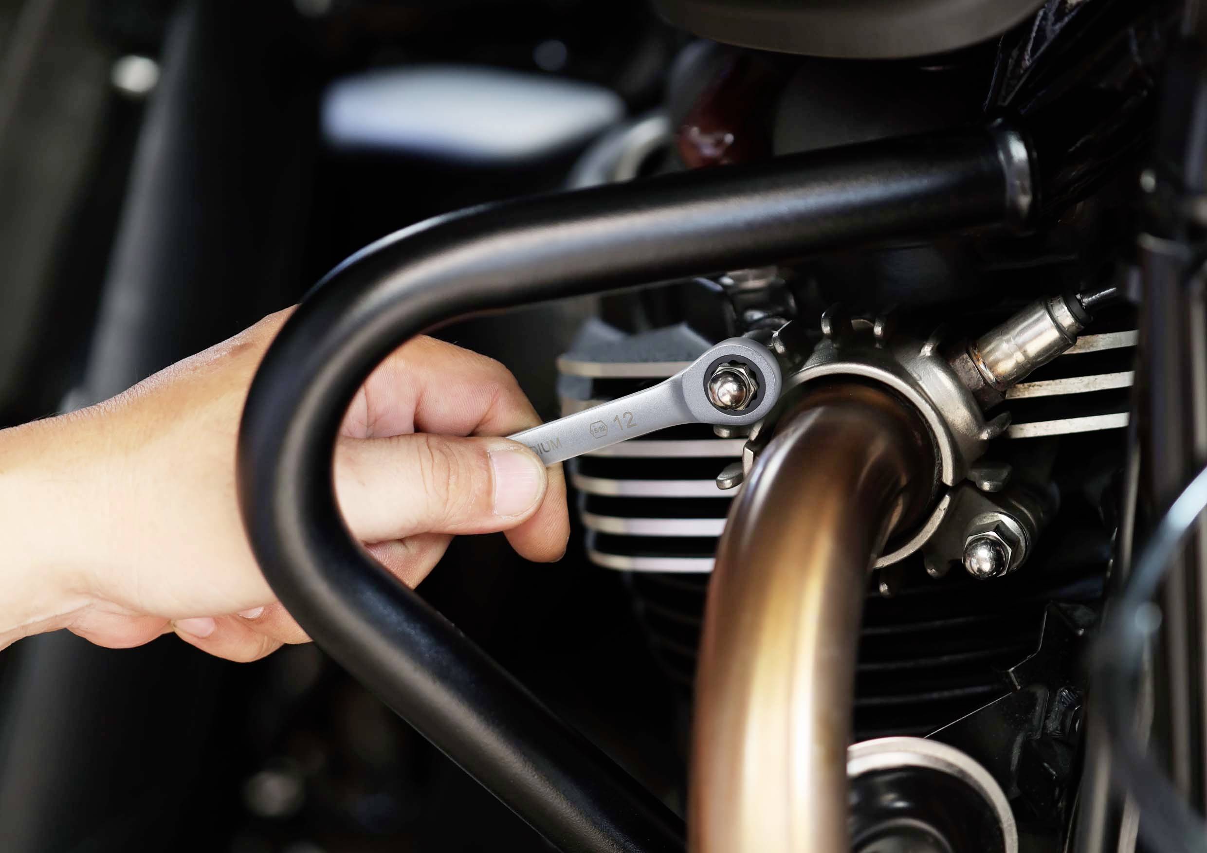 Eine Hand hält einen Schraubenschlüssel und zieht eine Schraube am Auspuff eines Motorrads fest.