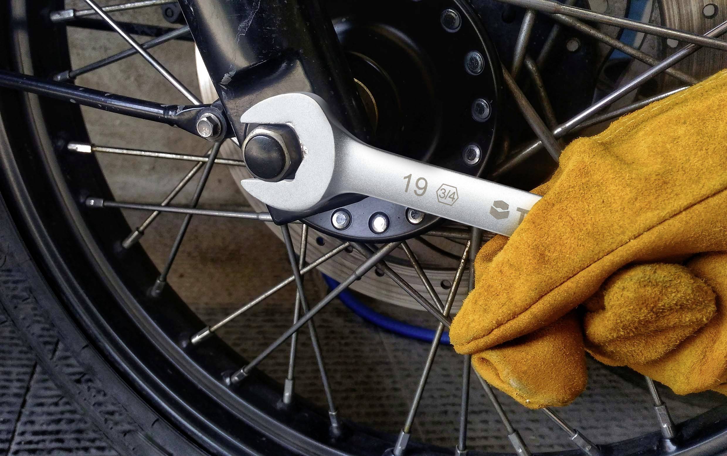 Eine Hand in gelbem Handschuh hält einen Schraubenschlüssel, der an einer Schraube eines Fahrrad- oder Motorrad-Rads angebracht ist.