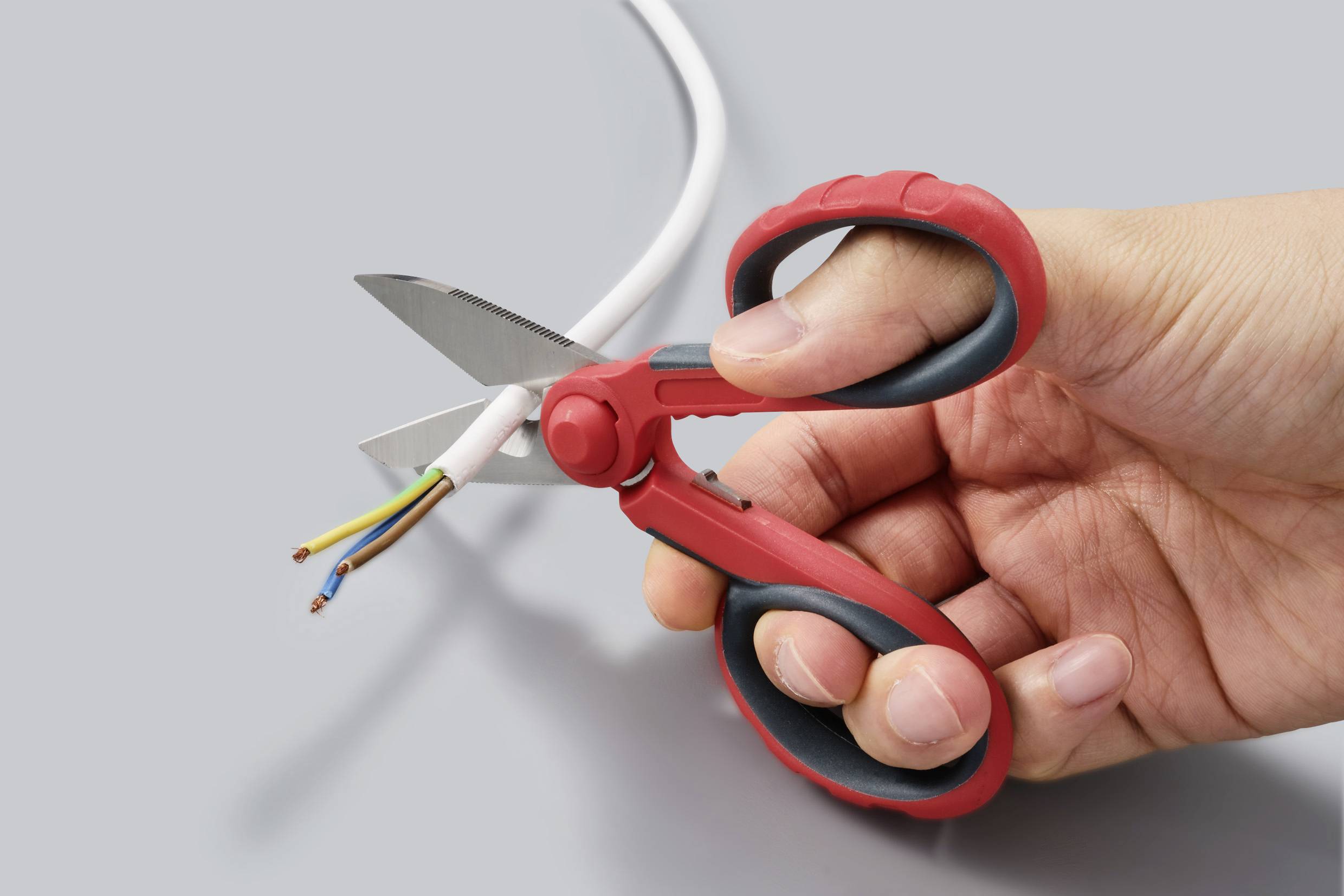 Hand mit roter Schere schneidet ein Kabel mit mehreren farbigen Drähten im Querschnitt.