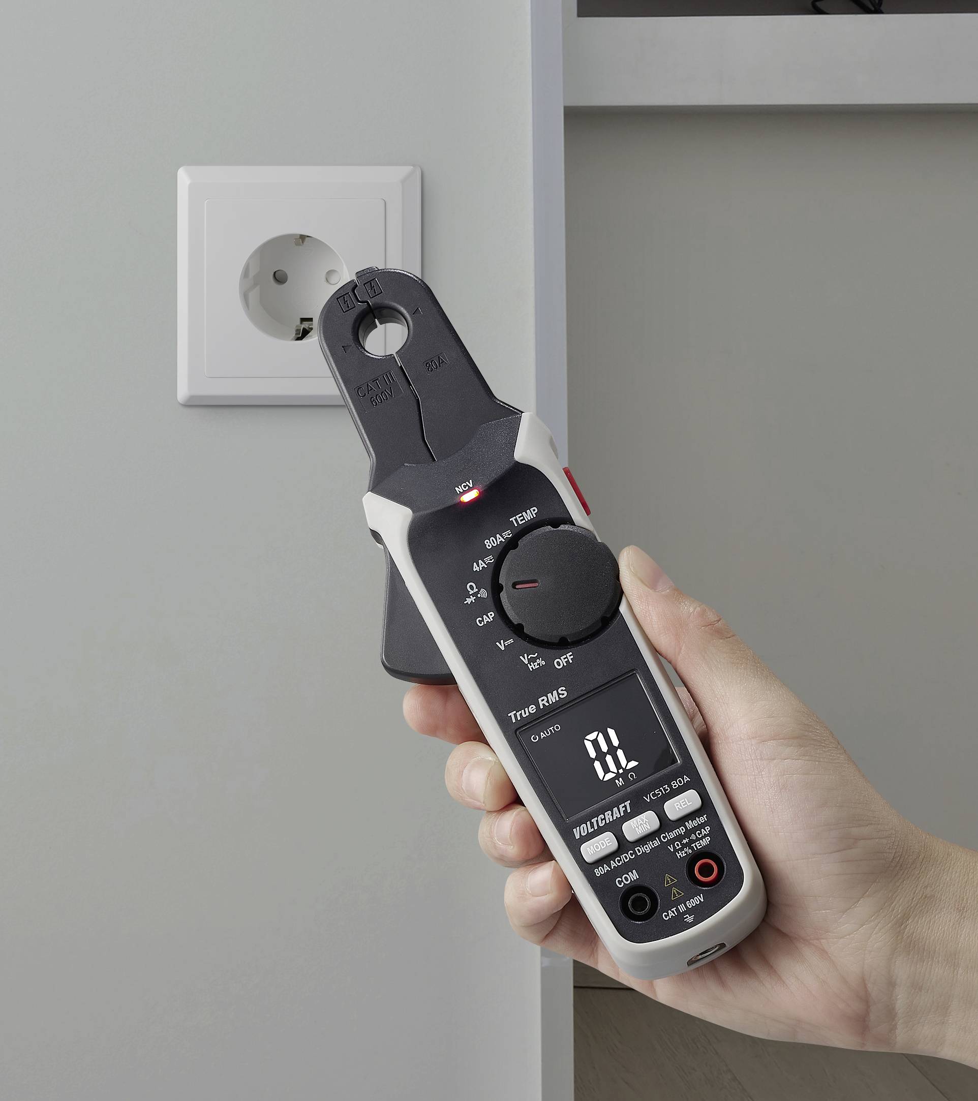 Eine Hand hält ein digitales Multimeter, das eine Steckdose misst. Das Display des Multimeters zeigt Messwerte an und deutet darauf hin, dass es gerade zur elektrischen Überprüfung verwendet wird.