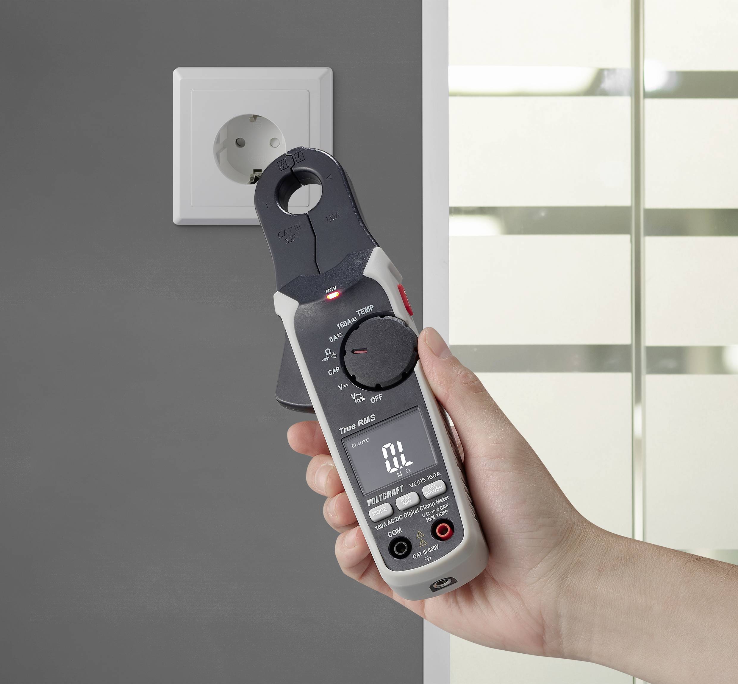 Eine Hand hält ein digitales Multimeter neben einer Steckdose an einer grauen Wand. Gerät zeigt '0 L' auf dem Display an.