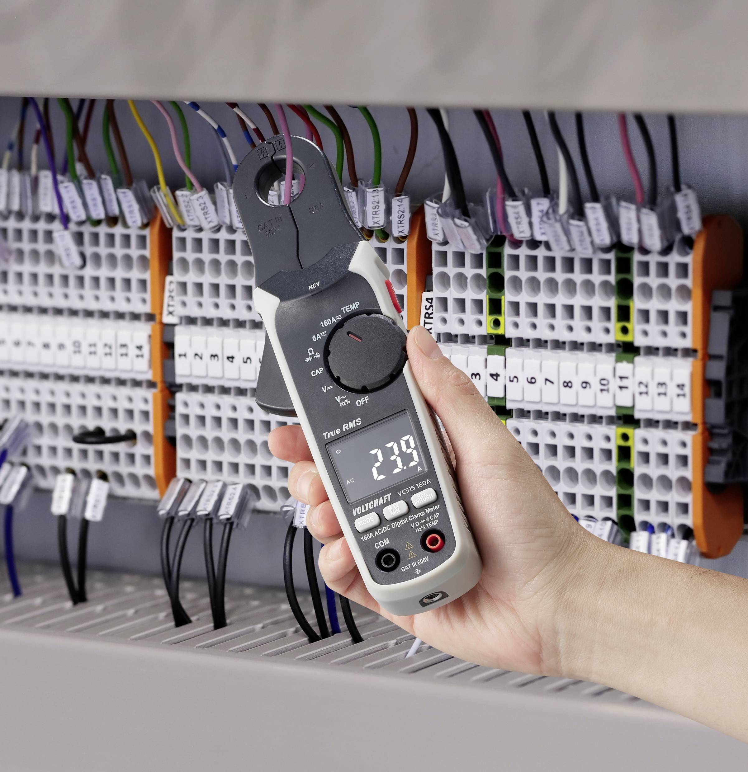 Eine Hand hält einen digitalen Stromzangenmesser vor einem Schaltkasten mit vielen Kabeln. Der Bildschirm zeigt '239'.