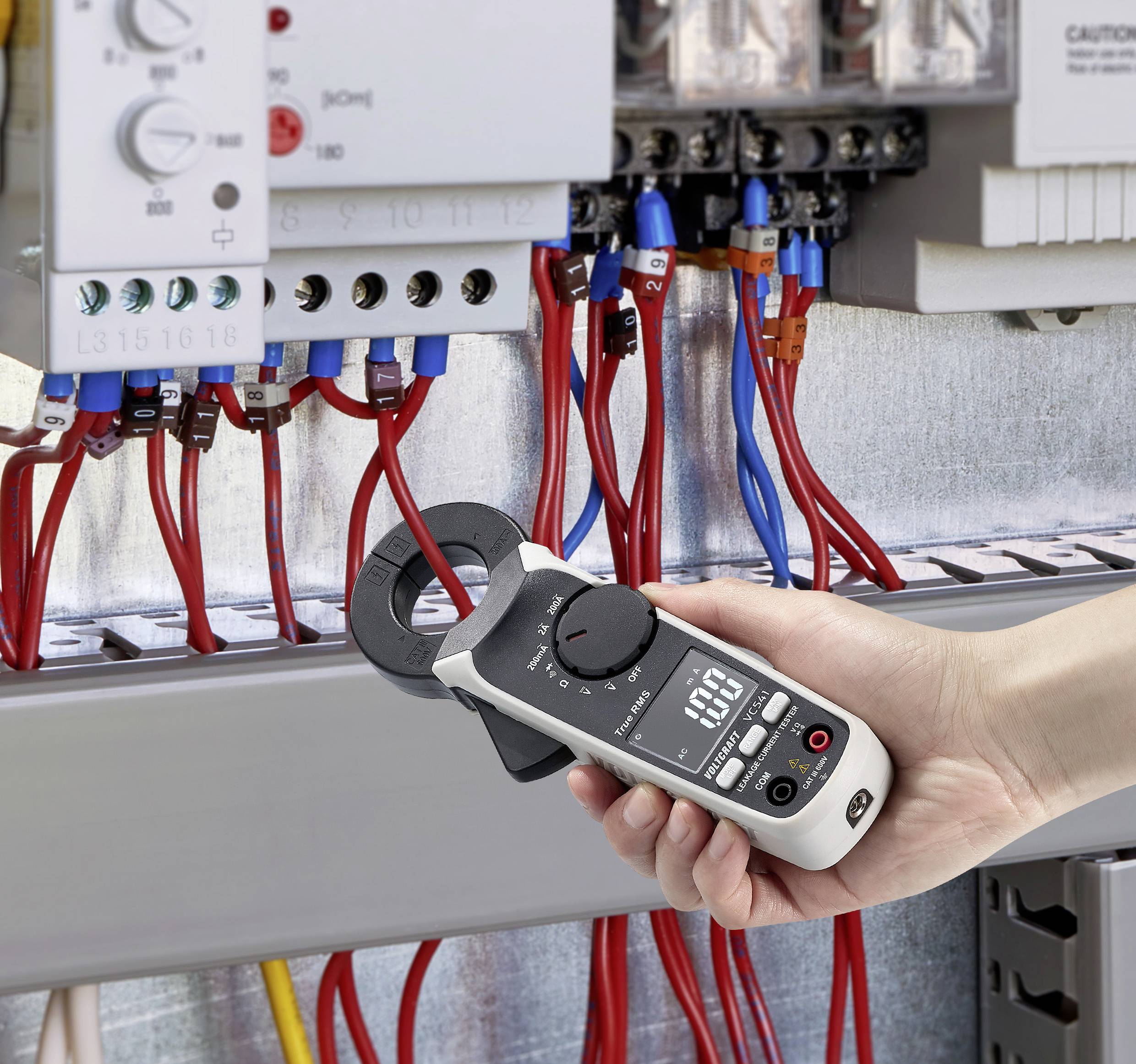 Eine Person hält ein digitales Multimeter vor einem offenen Schaltschrank, misst Stromwerte mit roten und blauen Anschlusskabeln.