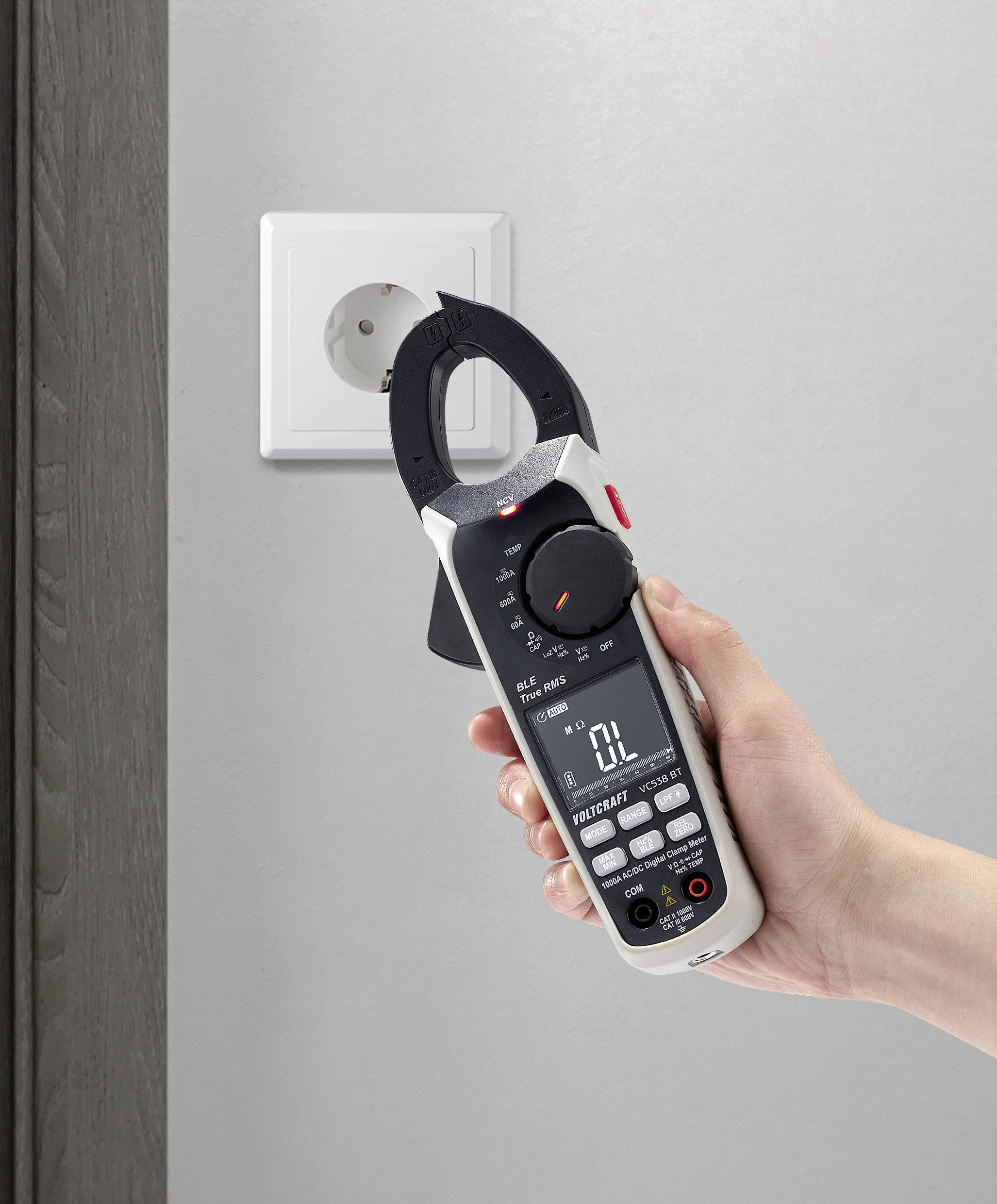 Eine Hand hält ein Multimeter vor eine Steckdose an der Wand. Das Multimeter zeigt elektrische Messwerte an.