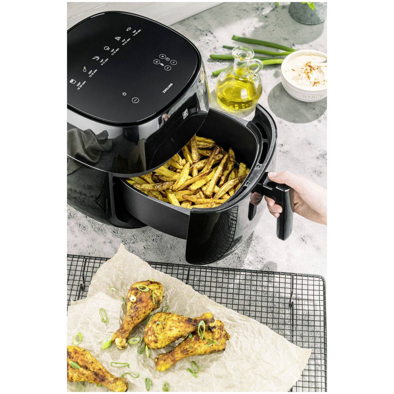 Eine Heißluftfritteuse mit geöffnetem Korb voller goldener Pommes Frites. Daneben liegt gebratenes Hähnchen auf einem Rost.