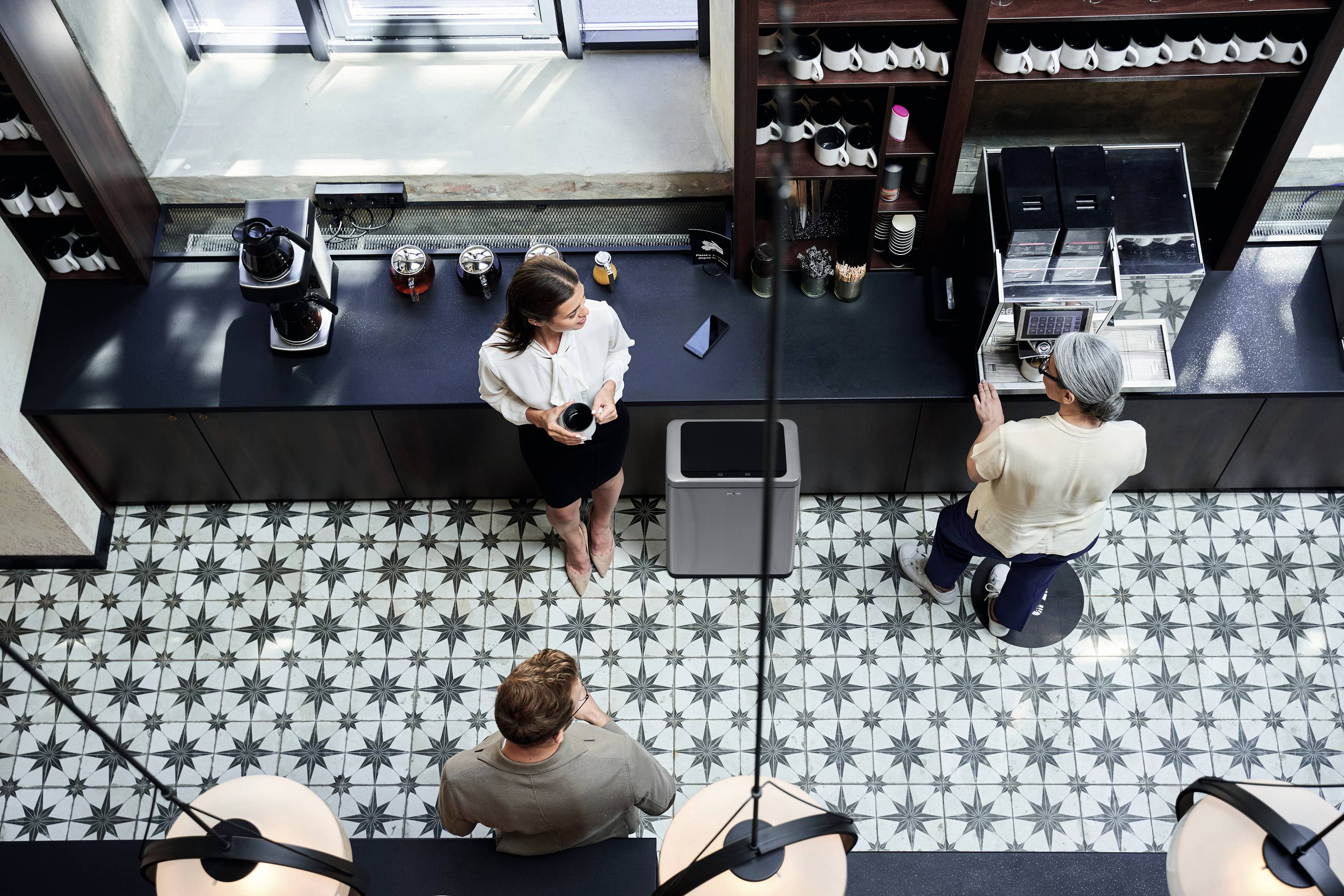 Zwei Frauen und ein Mann stehen in einem modernen Café an einer Theke. Eine Frau steht, die andere sitzt und bedient die Kaffeemaschine.