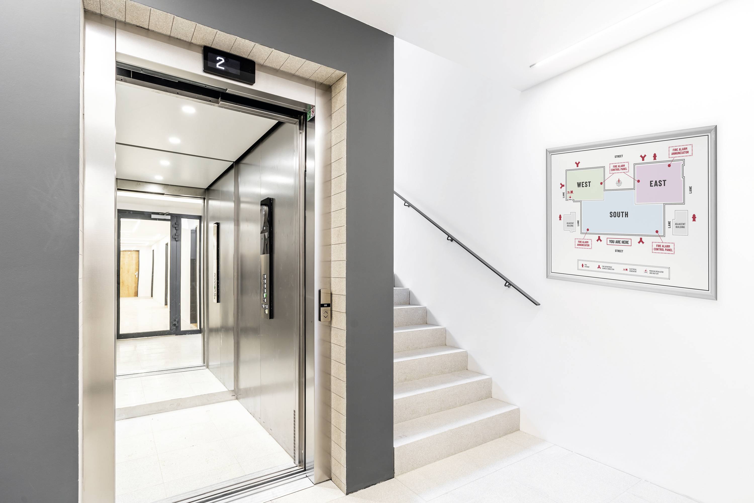 Offener Aufzug neben einer Treppe, mit Gebäudeplan an der Wand.