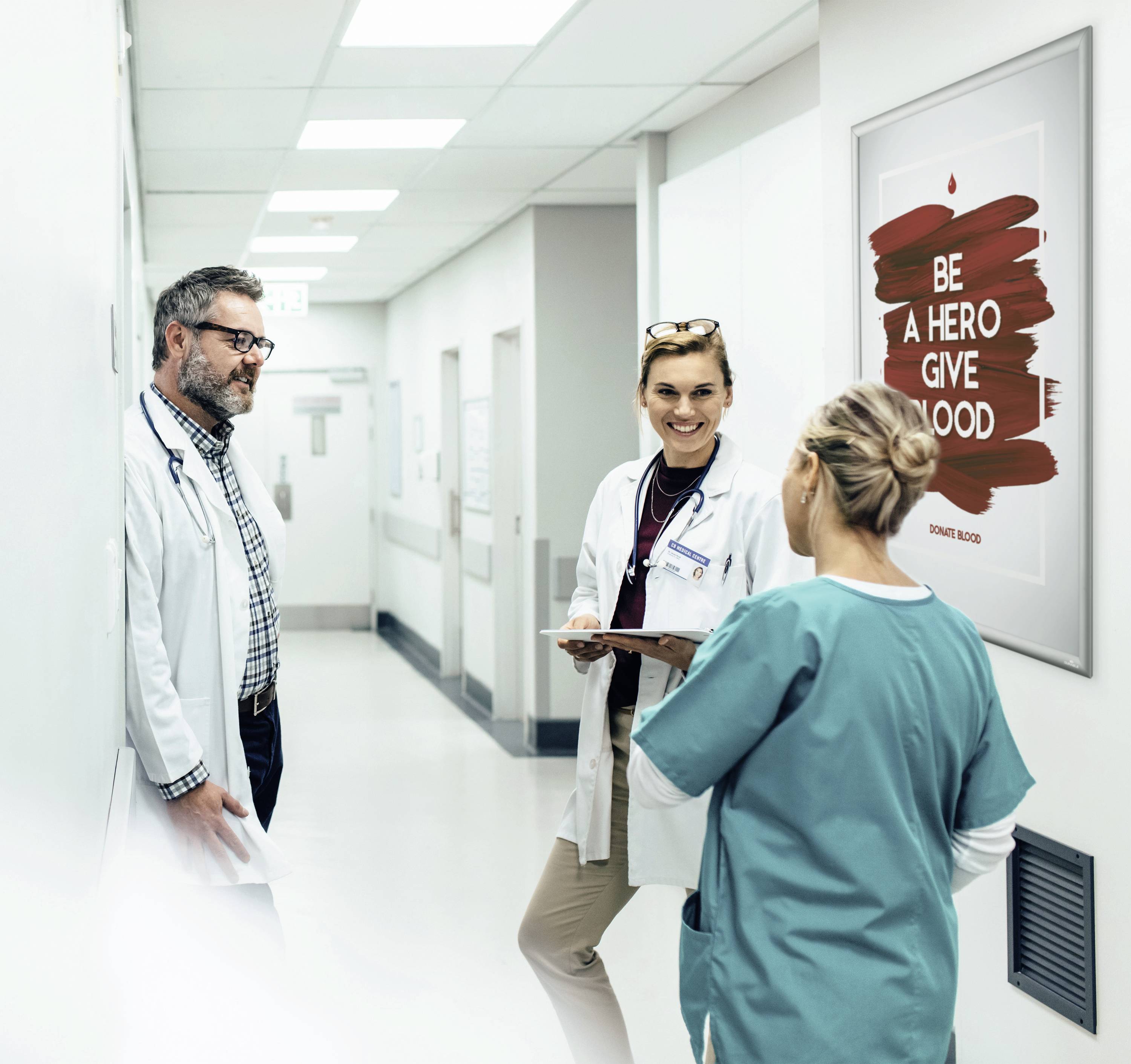 Drei medizinische Fachkräfte unterhalten sich im Krankenhausflur. Ein Plakat an der Wand sagt: 'Sei ein Held, spende Blut'.