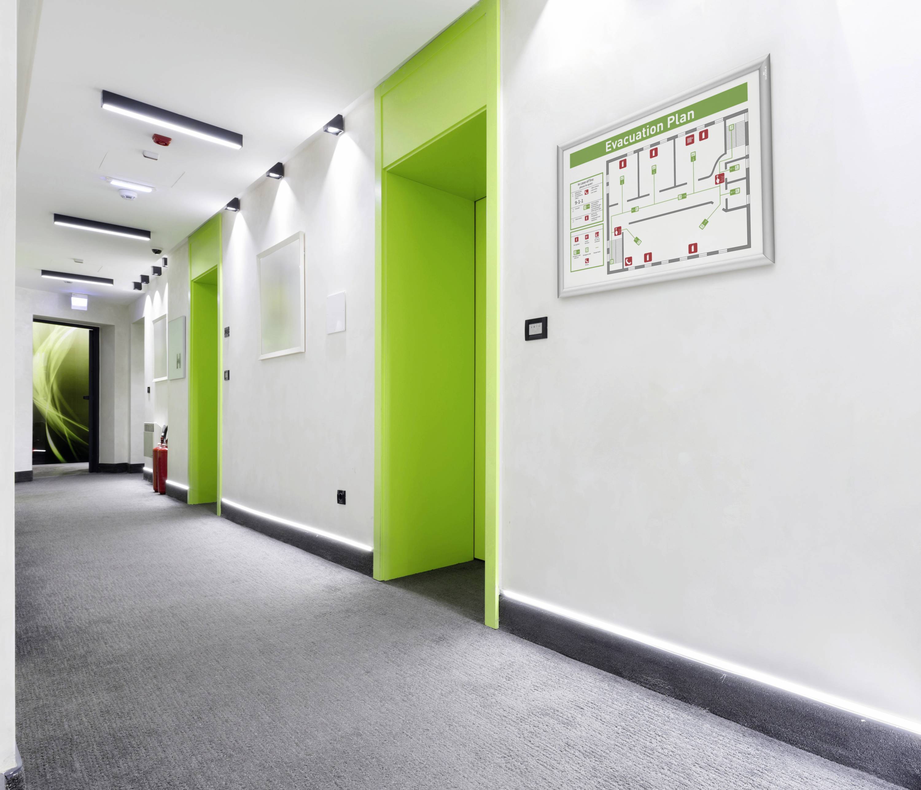 Ein heller Flur mit grün gestrichenen Türen und einem Evakuierungsplan an der Wand. Links hinten steht ein Feuerlöscher.