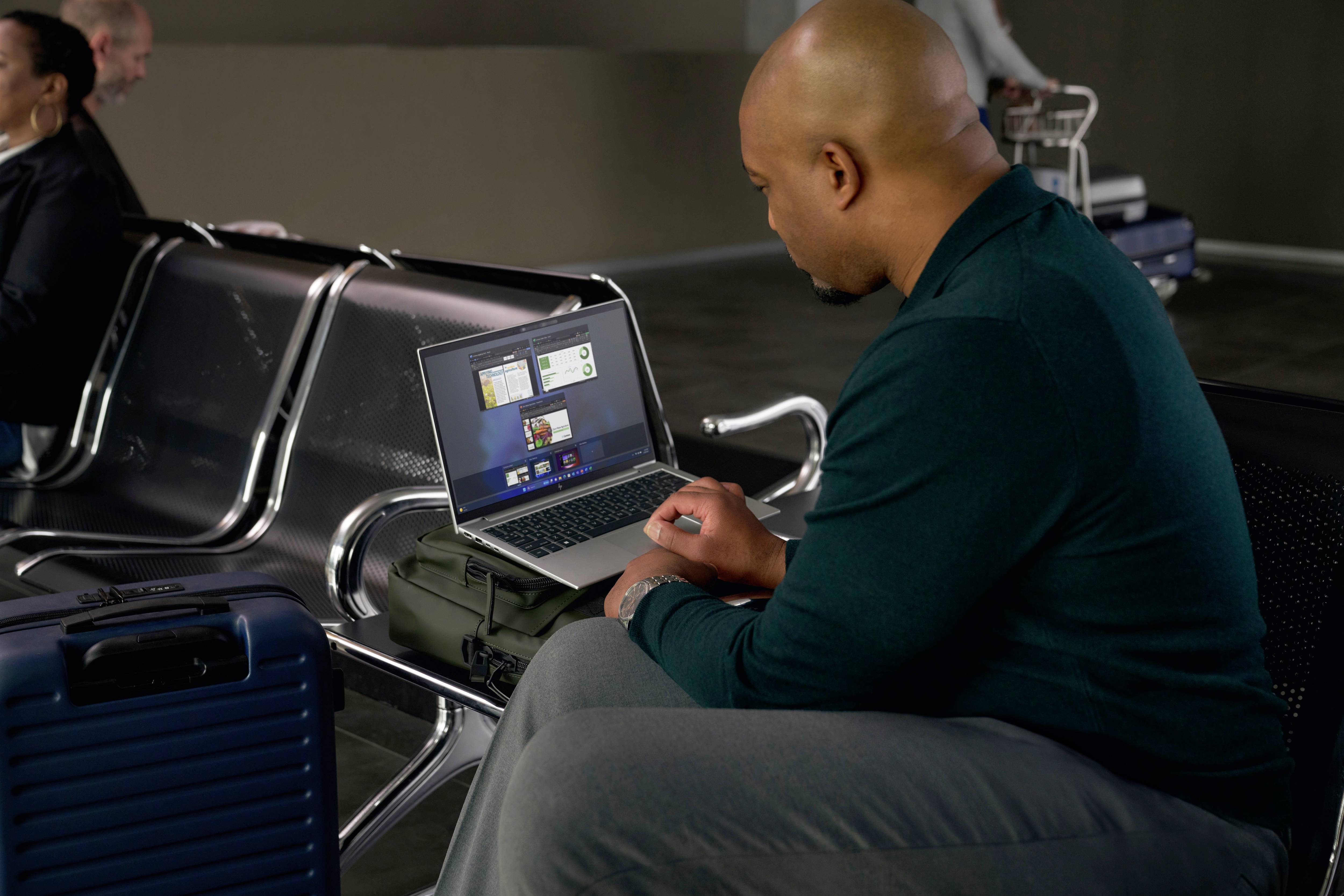 Ein Mann arbeitet an einem Laptop in einem Wartebereich mit Gepäck neben sich. Im Hintergrund sind weitere Personen und Gepäckwagen zu sehen.