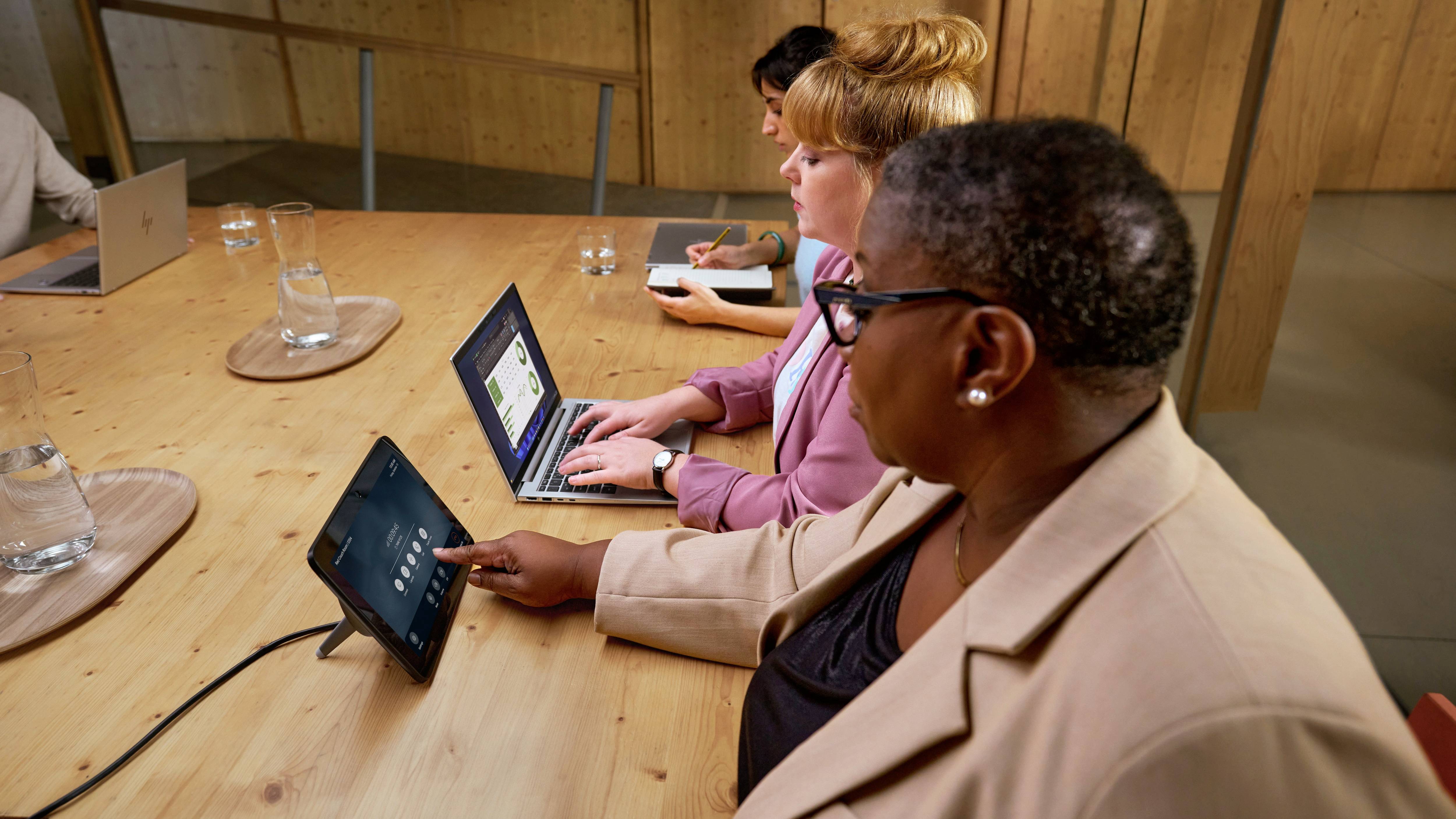 Drei Personen sitzen an einem Konferenztisch mit Laptops. Eine Person bedient ein Tablet. Alle konzentrieren sich auf ihre Geräte.