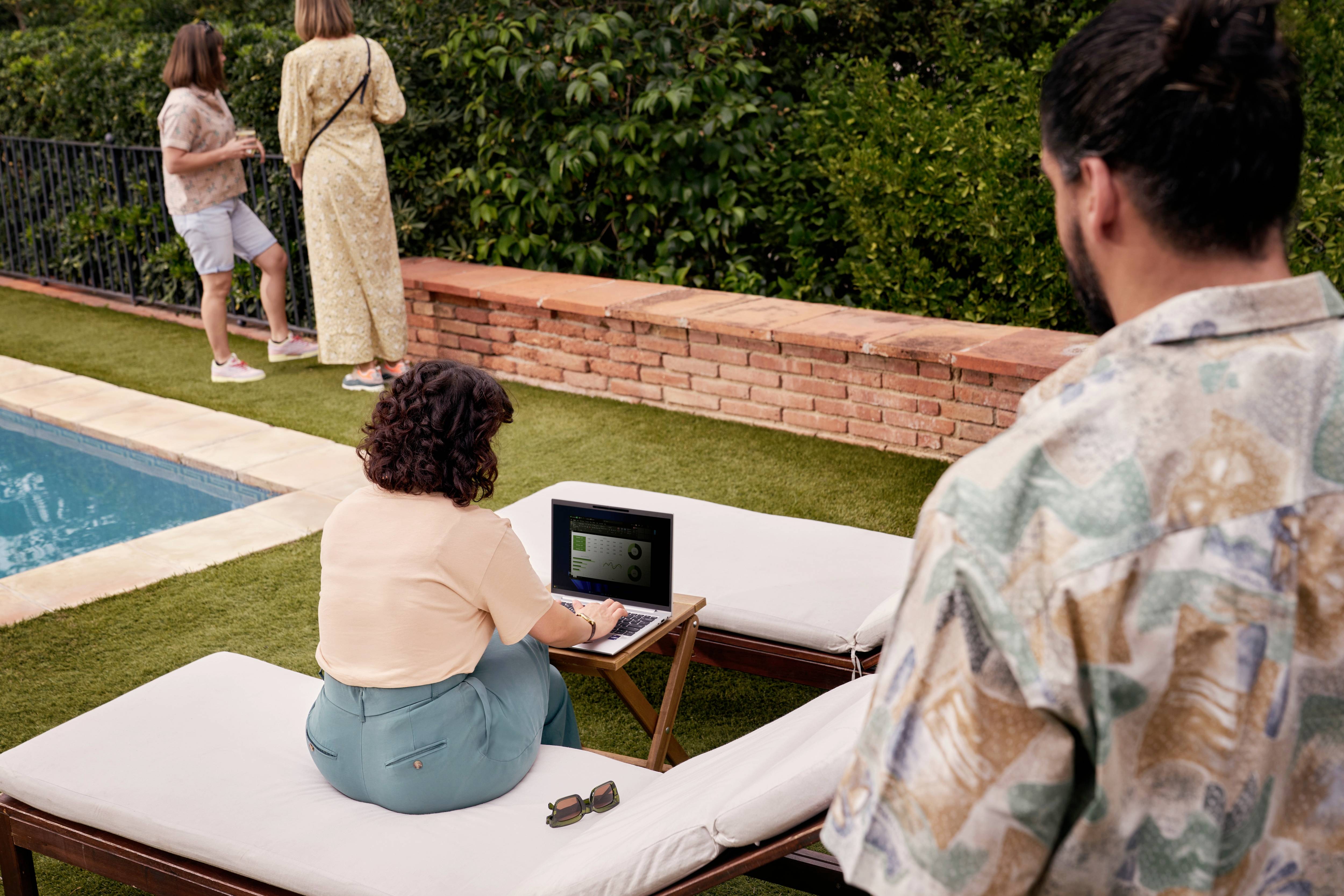 Eine Person arbeitet an einem Laptop auf einer Liege am Pool, während zwei weitere Personen im Hintergrund stehen, umgeben von Bäumen.