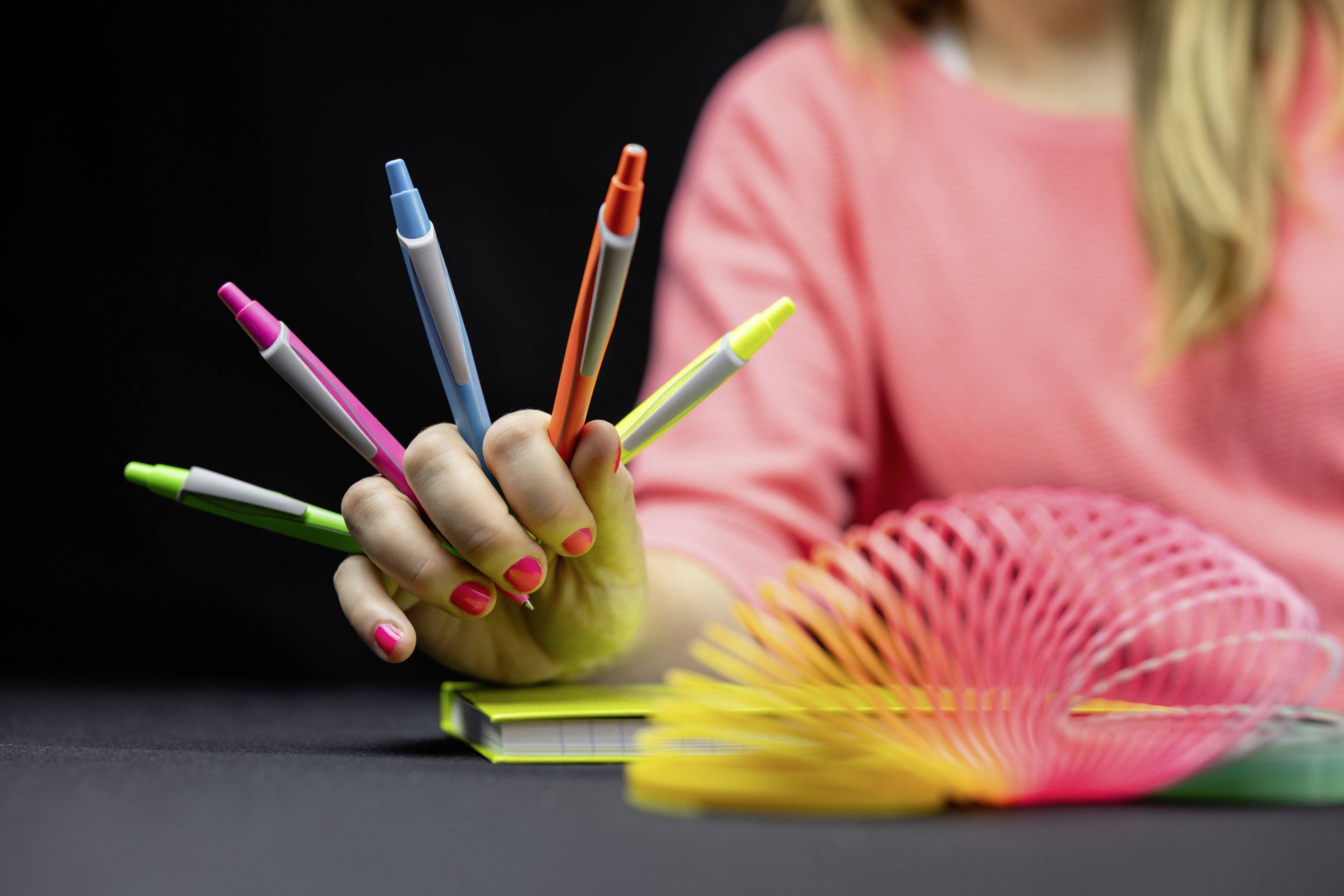 Eine Person hält fünf bunte Stifte zwischen den Fingern in einer verspielten Art. Im Vordergrund ist ein bunter Spiral-Slinky zu sehen.