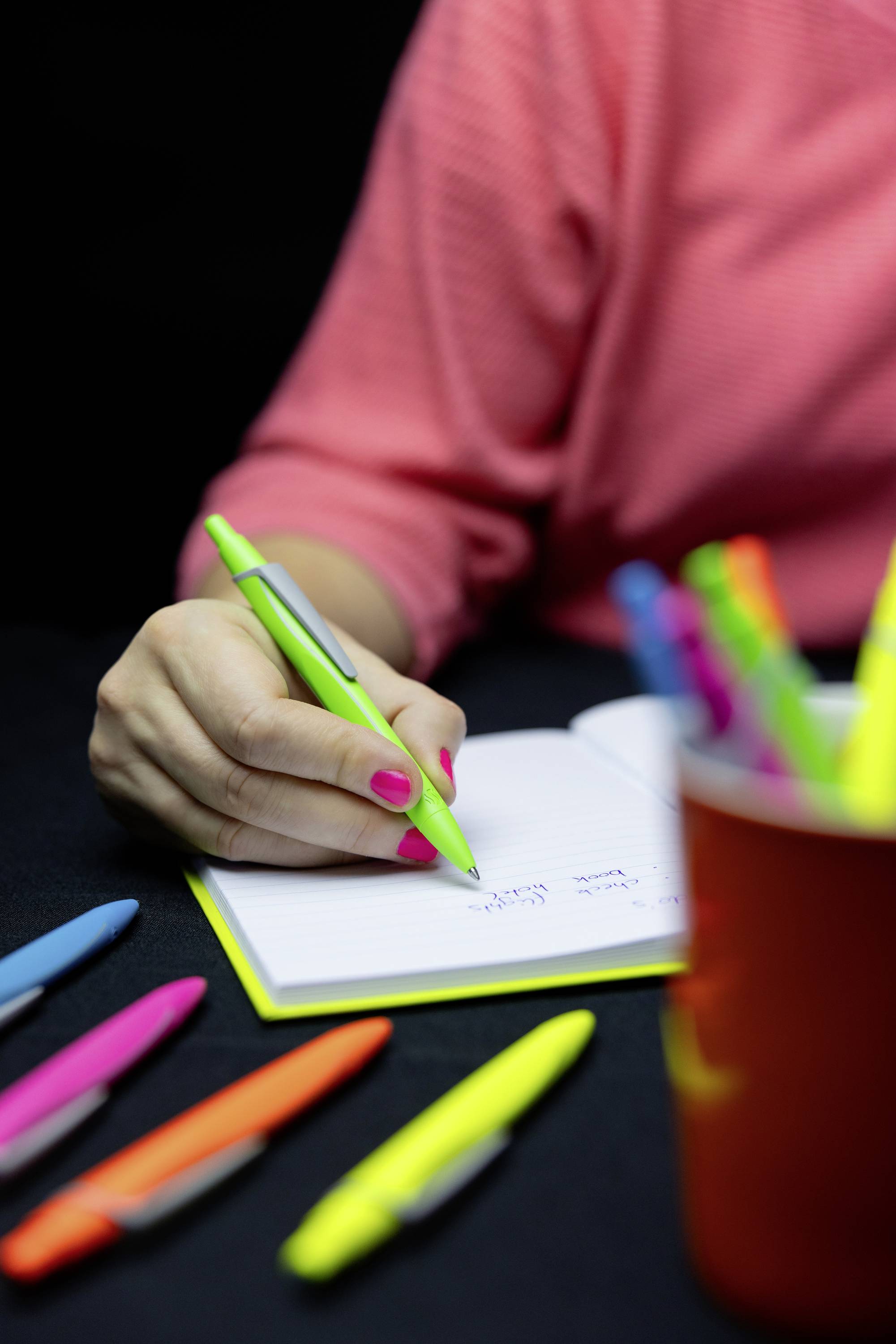 Eine Person schreibt mit einem grünen Stift in ein Notizbuch. Im Vordergrund liegen bunte Stifte verstreut, und ein Becher mit weiteren Stiften ist teilweise sichtbar.