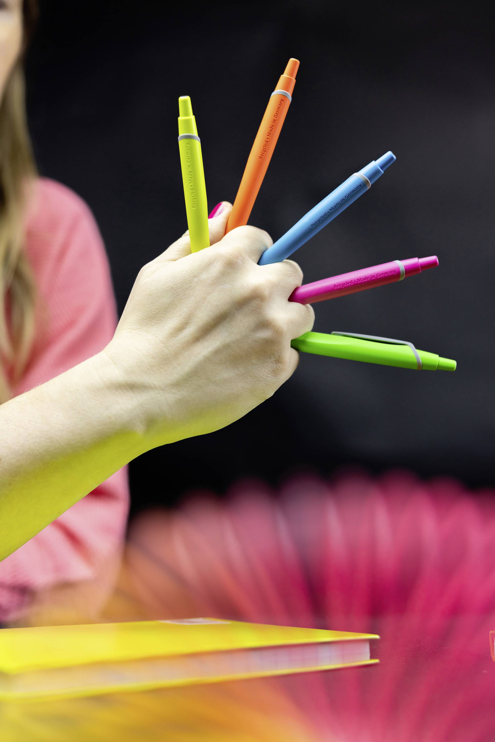 Hand hält fünf bunte Stifte, die wie ein Fächer angeordnet sind; unscharfer Hintergrund mit rotem und schwarzem Farbverlauf.
