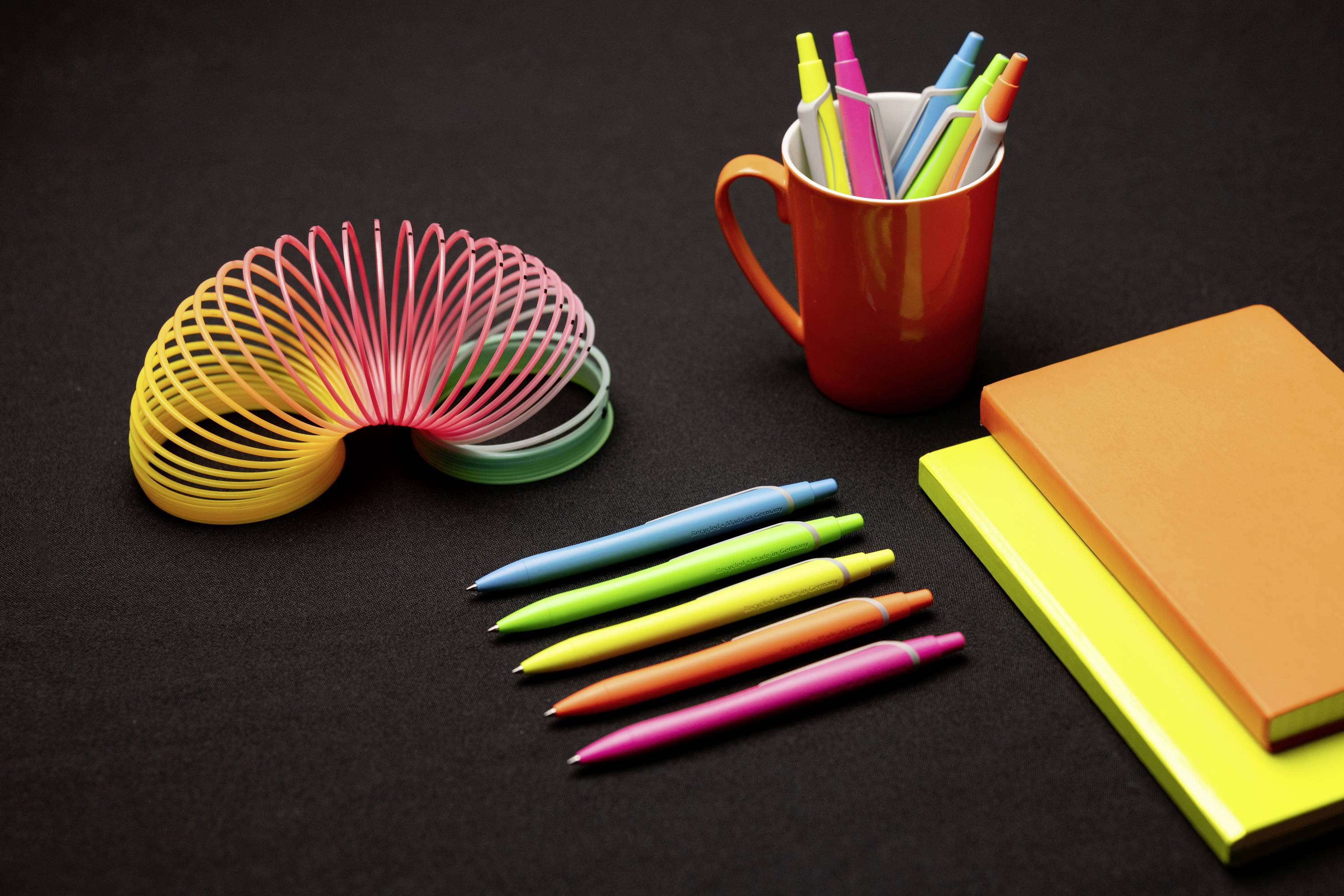 Bunte Büromaterialien auf schwarzem Hintergrund: Stifte, ein rotes Tasse mit Markern, ein Regenbogen-Slinky, zwei Notizblöcke.
