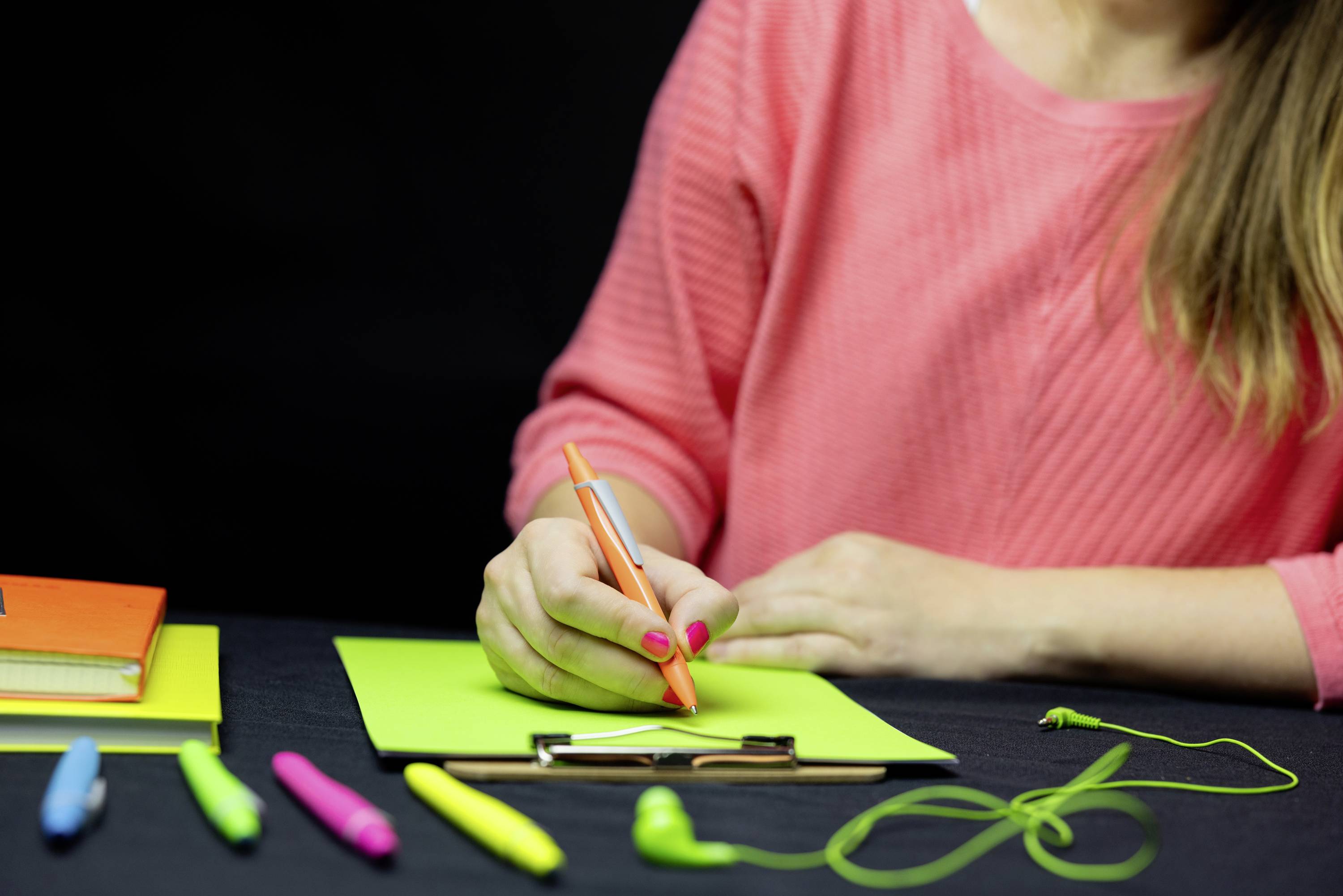 Eine Person in einem rosa Oberteil schreibt mit einem orangefarbenen Stift auf hellgrünem Papier. Verschiedene Stifte liegen auf dem Tisch.