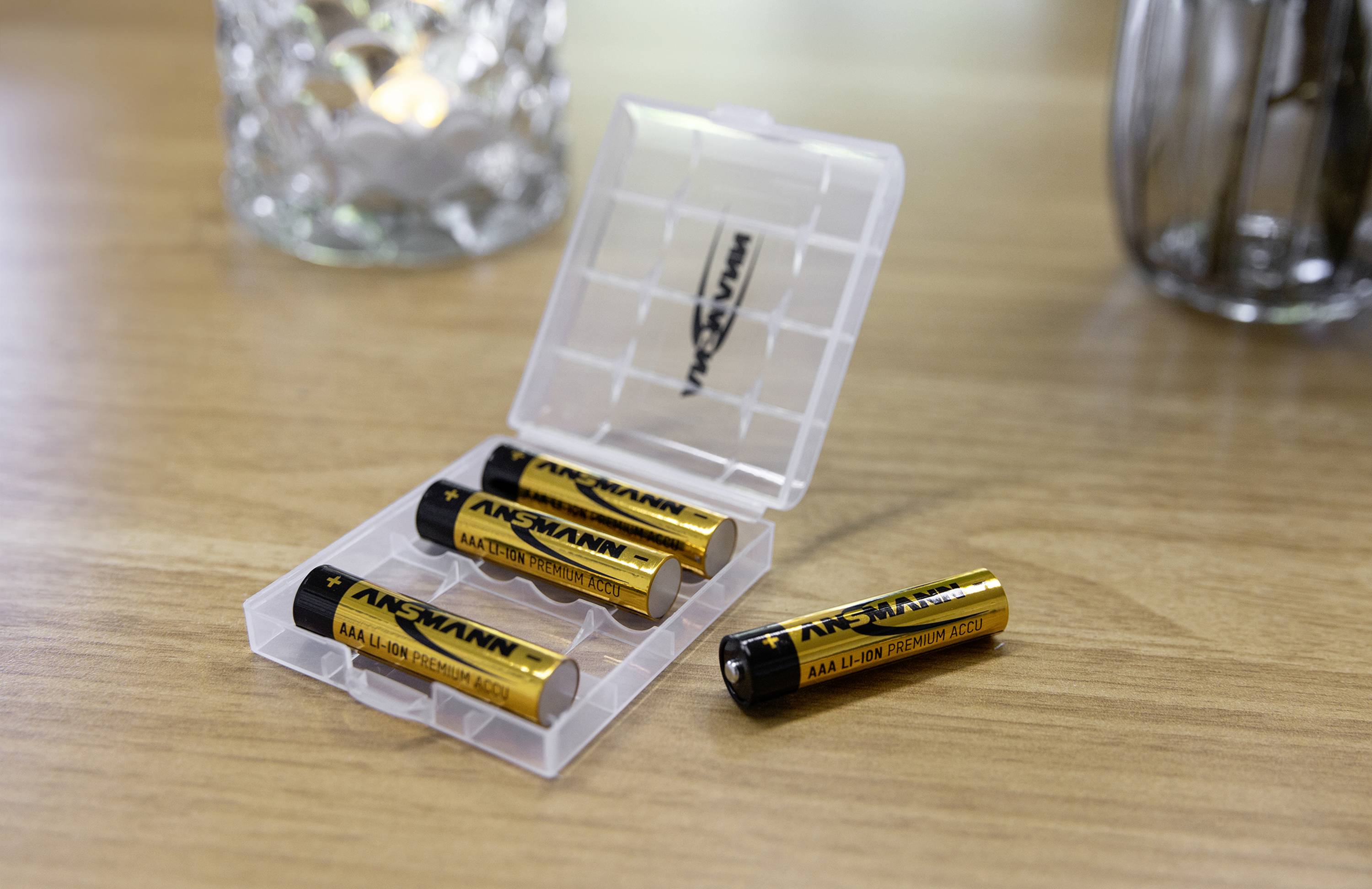 Vier AA-Batterien in einer transparenten Box auf einem Holztisch; eine Batterie liegt daneben.