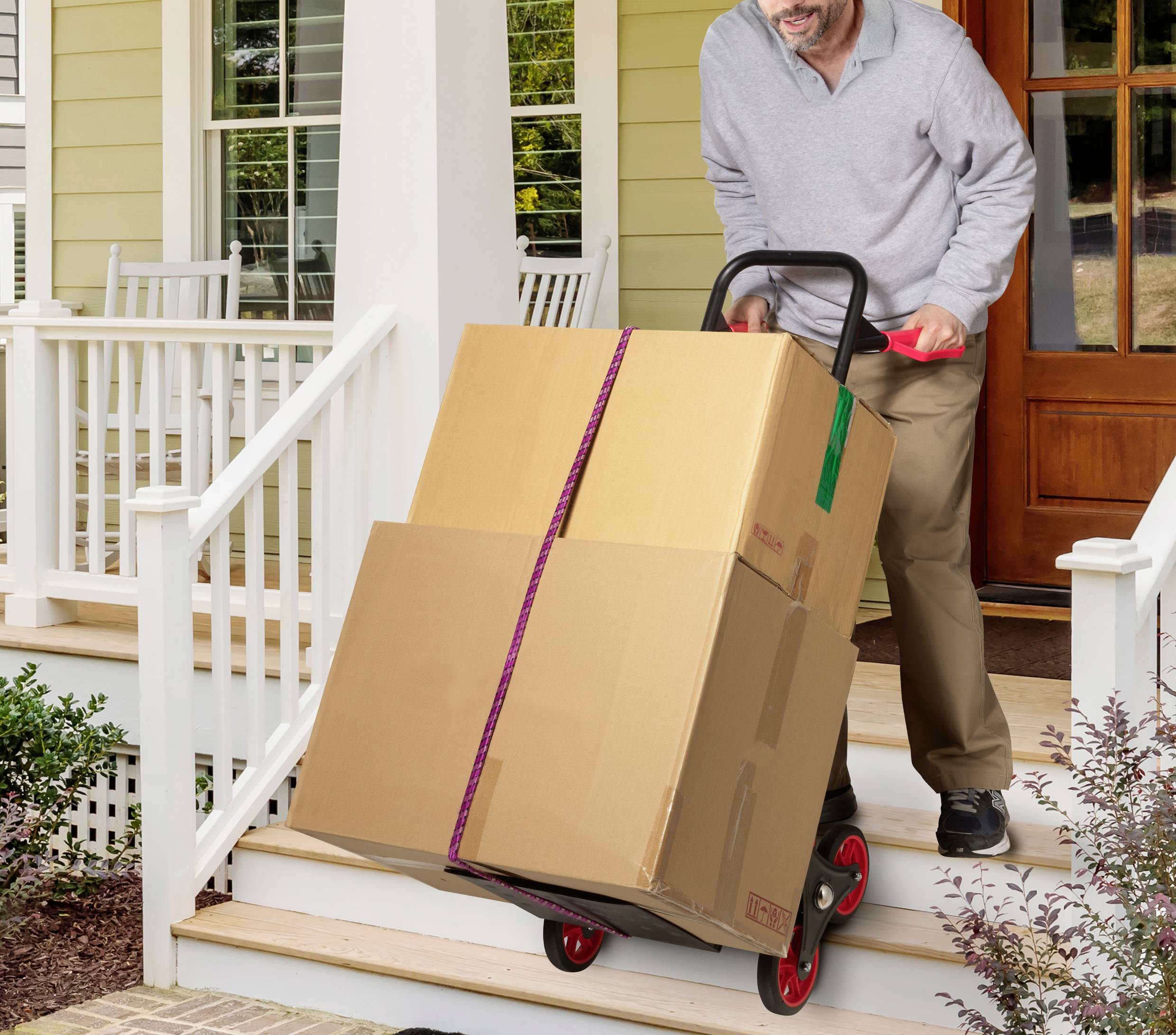 Ein Mann transportiert auf einem Trolley zwei große Kartons die Stufen einer Veranda hinauf. Im Hintergrund ist ein Haus erkennbar.