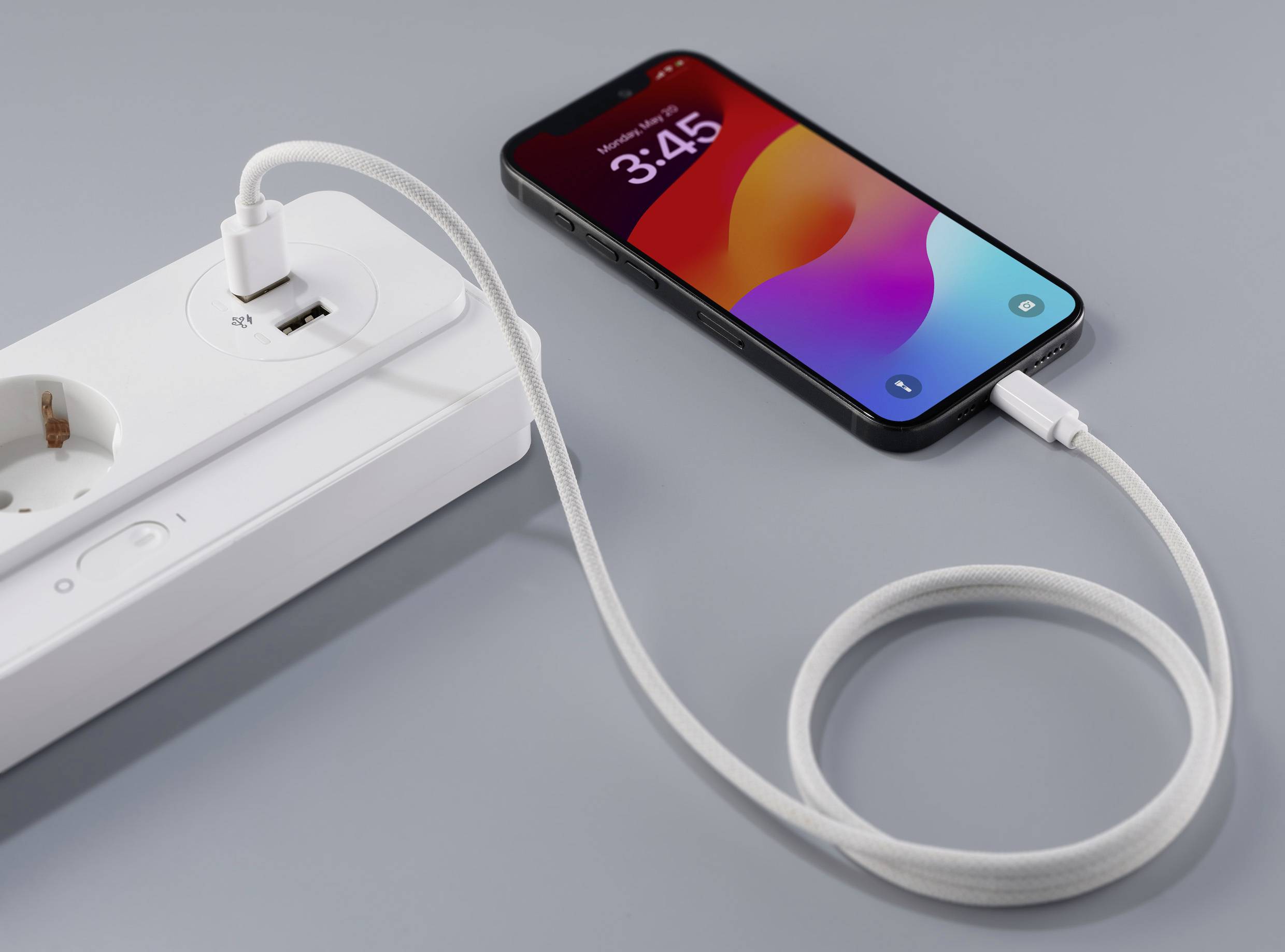 Smartphone mit Ladesymbol ist über ein USB-Kabel mit einer Steckdosenleiste verbunden. Das Display zeigt die Uhrzeit 3:45.