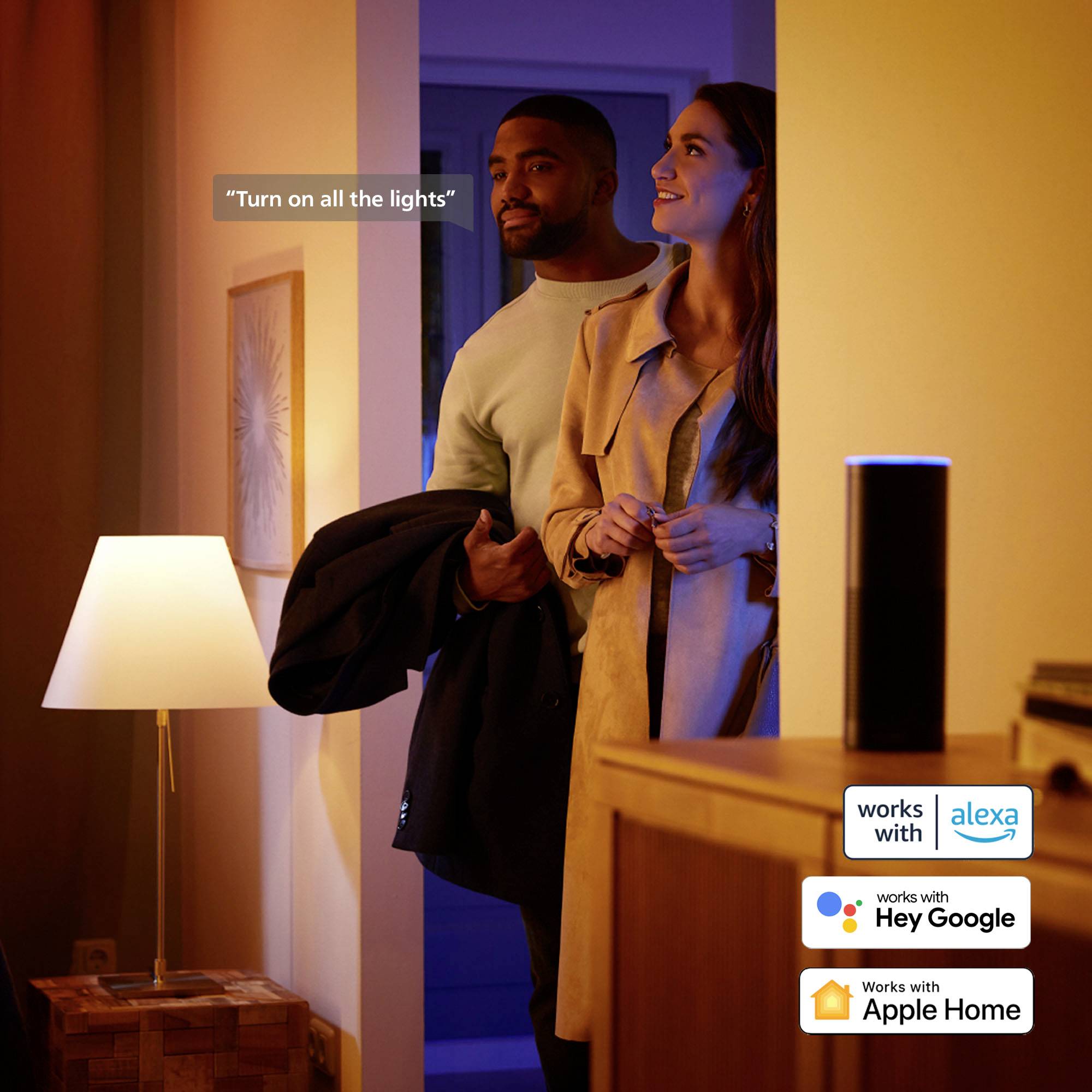 Ein Paar betritt ein Zimmer, das von Lampenlicht erhellt wird. Ein smarter Lautsprecher ist aktiv, mit Symbolen für Alexa, Hey Google und Apple Home.
