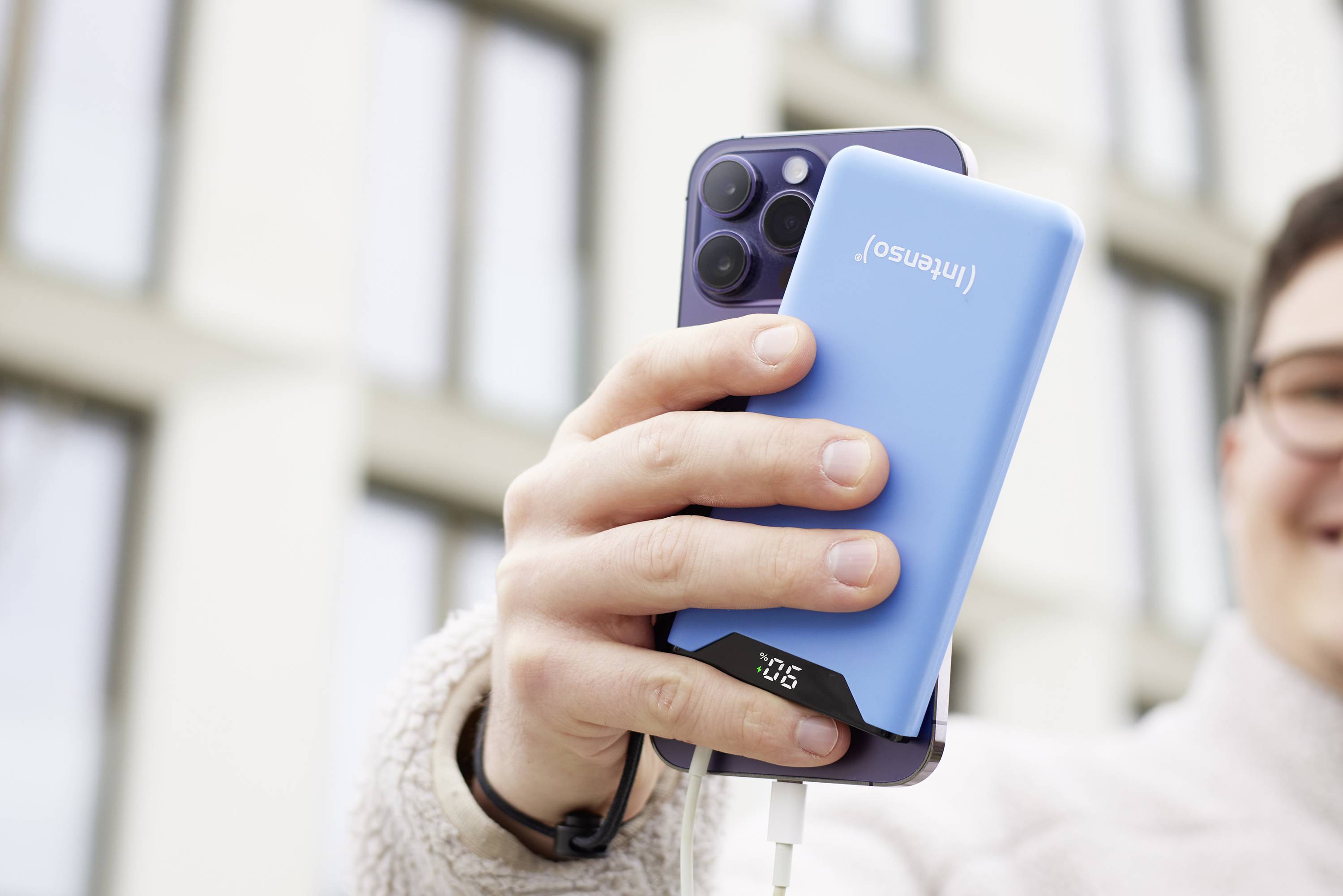 Person hält ein Smartphone und eine blaue Powerbank, die ein Kabel angeschlossen hat, in der Hand. Gebäude im Hintergrund.