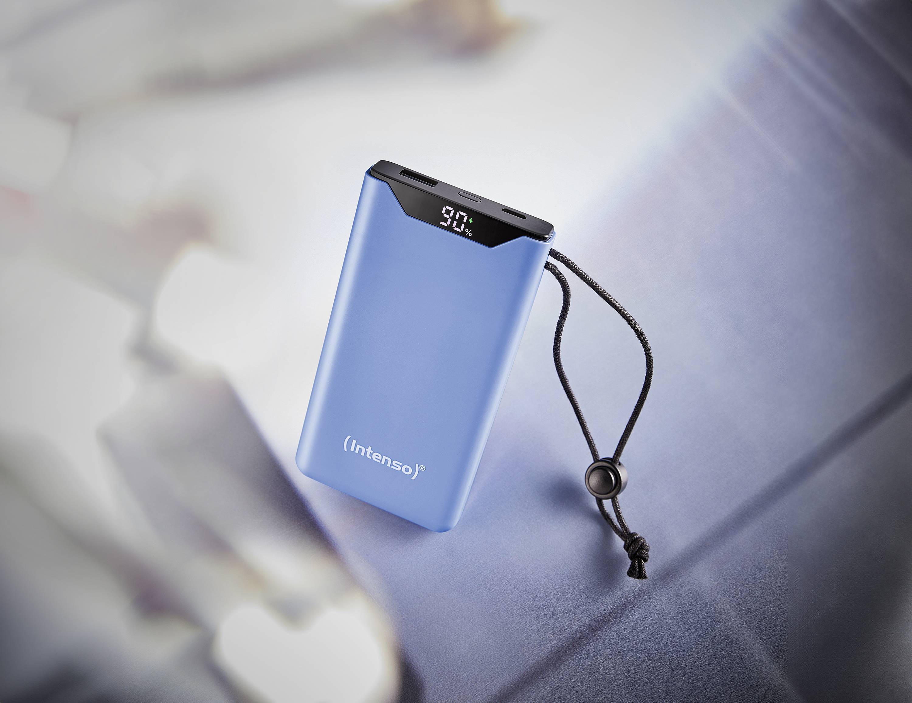 Blaue Powerbank mit schwarzem Band, digitales Display zeigt '90%' an. Liegt auf einer unscharfen, reflektierenden Oberfläche.